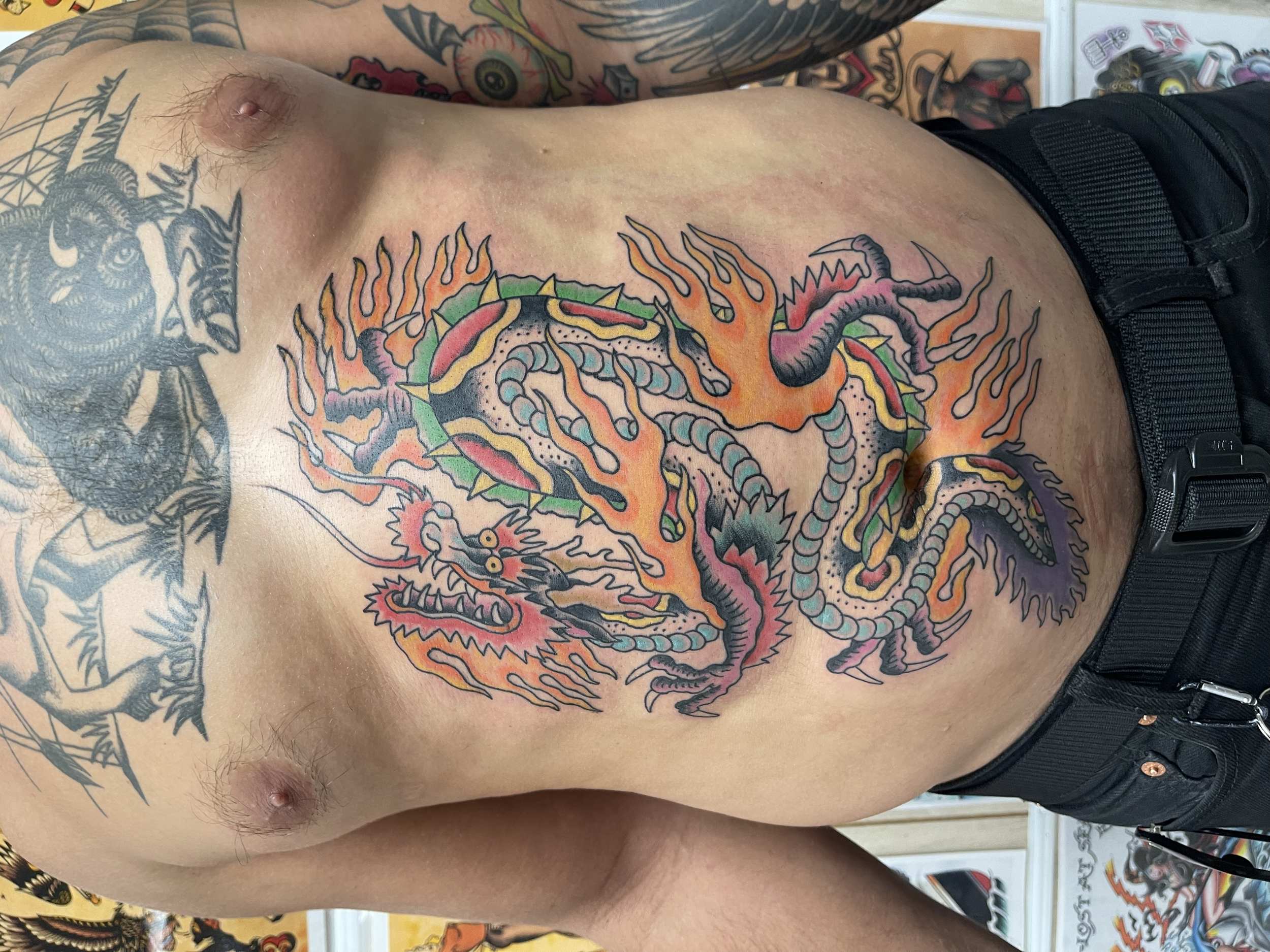 A person's torso with colorful tattoos, including a dragon, a snake with flames, and other designs on the chest and arm.