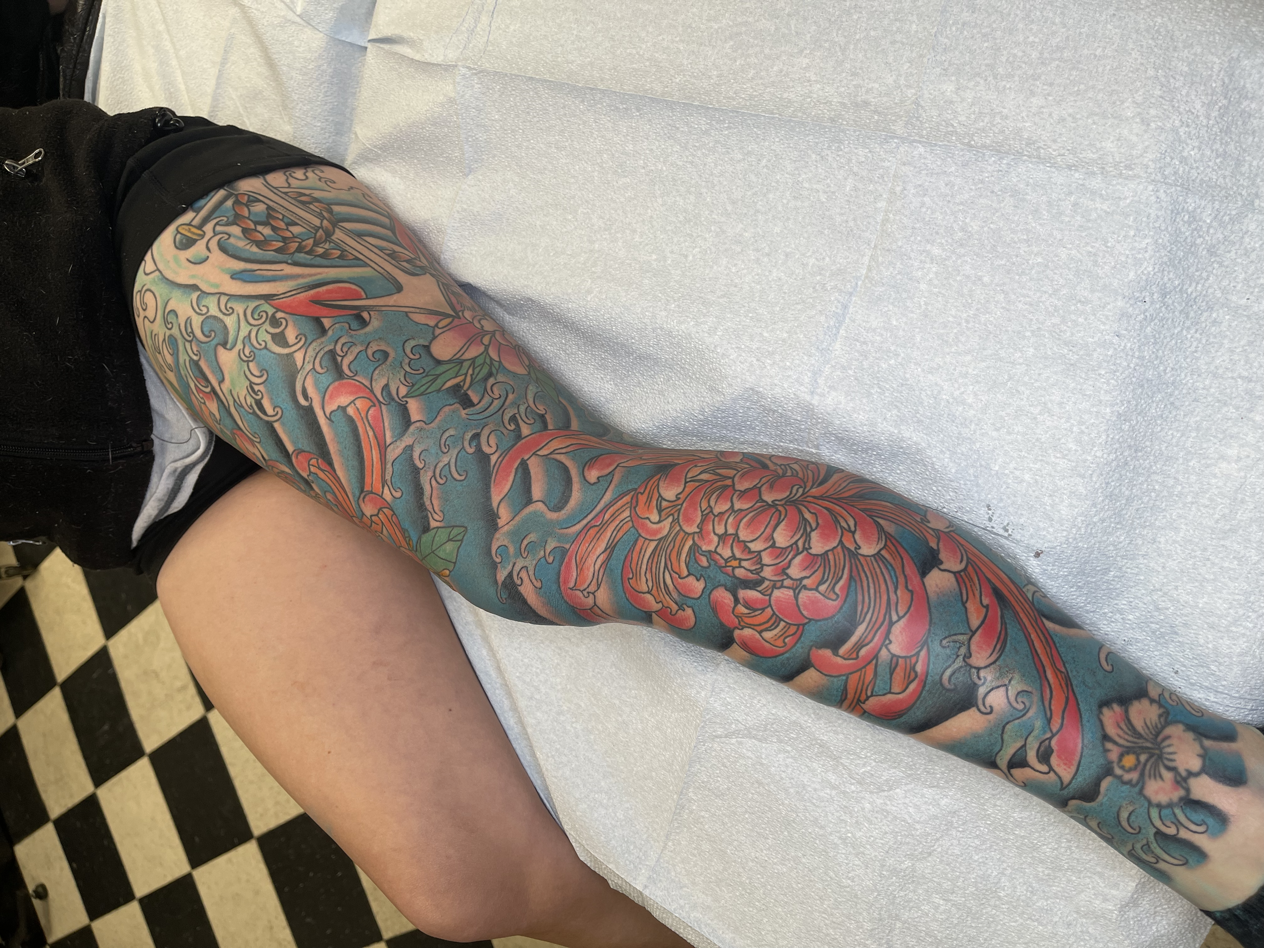 Colored tattoo sleeve featuring waves, a dragon, and flowers on a person's arm, lying on a white medical table with black-and-white checkered flooring visible in the background.