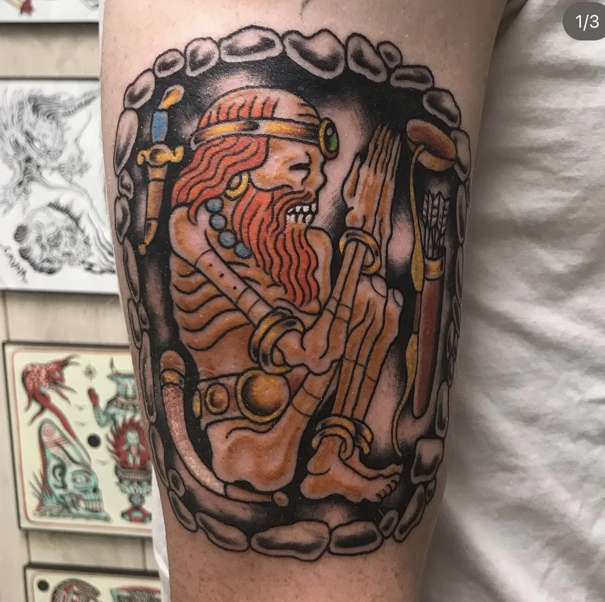Tattoo of a Viking warrior with long red hair and beard, wearing a helmet with a green gem, holding a sword and shield, surrounded by a stone border.