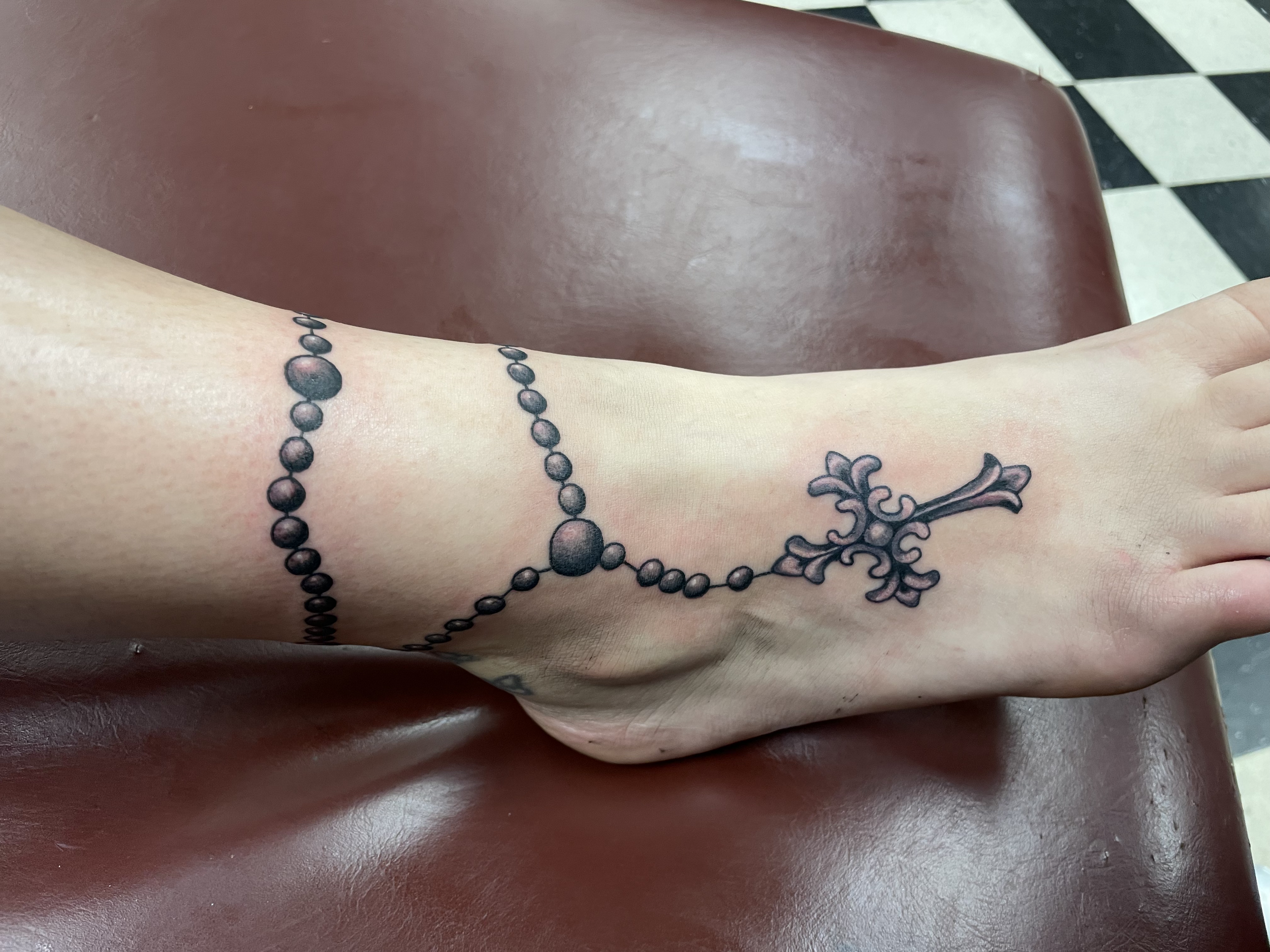 A close-up of a person's arm with a tattoo of a string of beads and a decorative floral design, resting on a red chair, with a black and white checkered floor in the background.