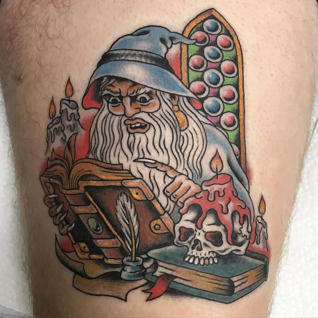 Tattoo of a wizard with a long beard, wearing a hat, reading a book, with candles, a skull, and a stained-glass window in the background.