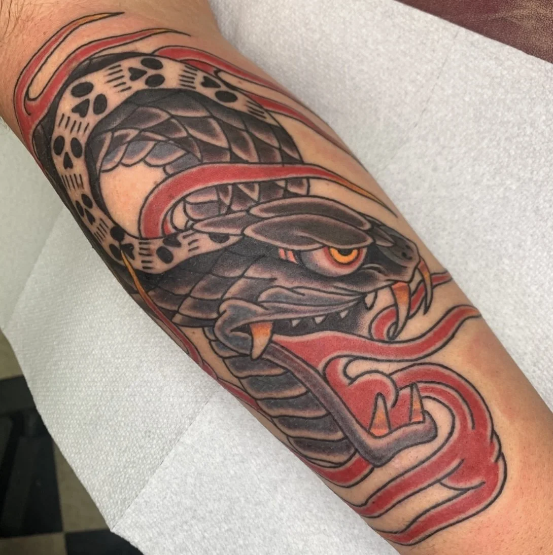 Close-up of a colorful tattoo on a person's arm featuring a fierce black snake with red accents and yellow eyes, coiled with its mouth open and fangs visible.