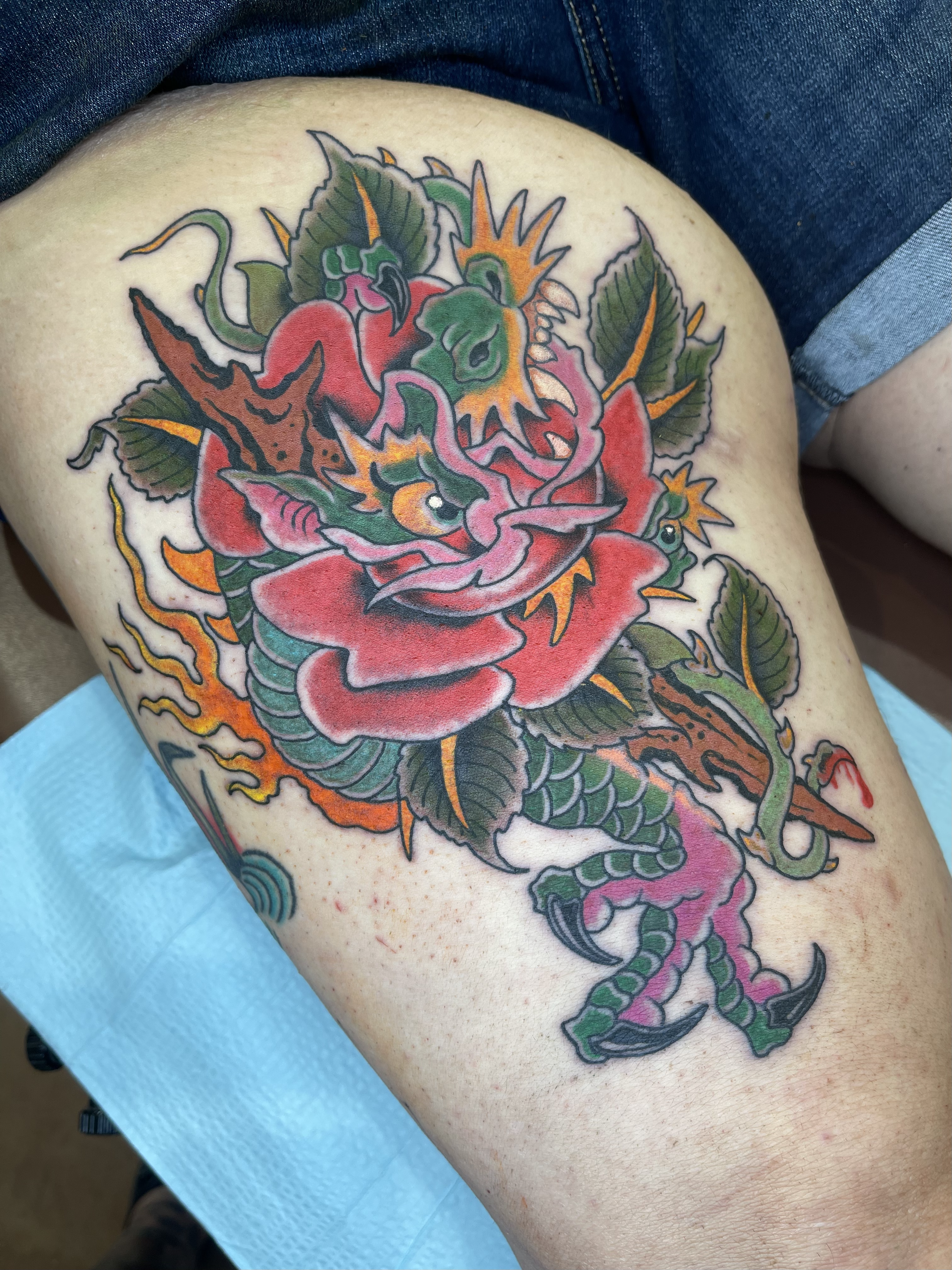 Colorful tattoo of a dragon intertwined with a rose on a person's thigh.