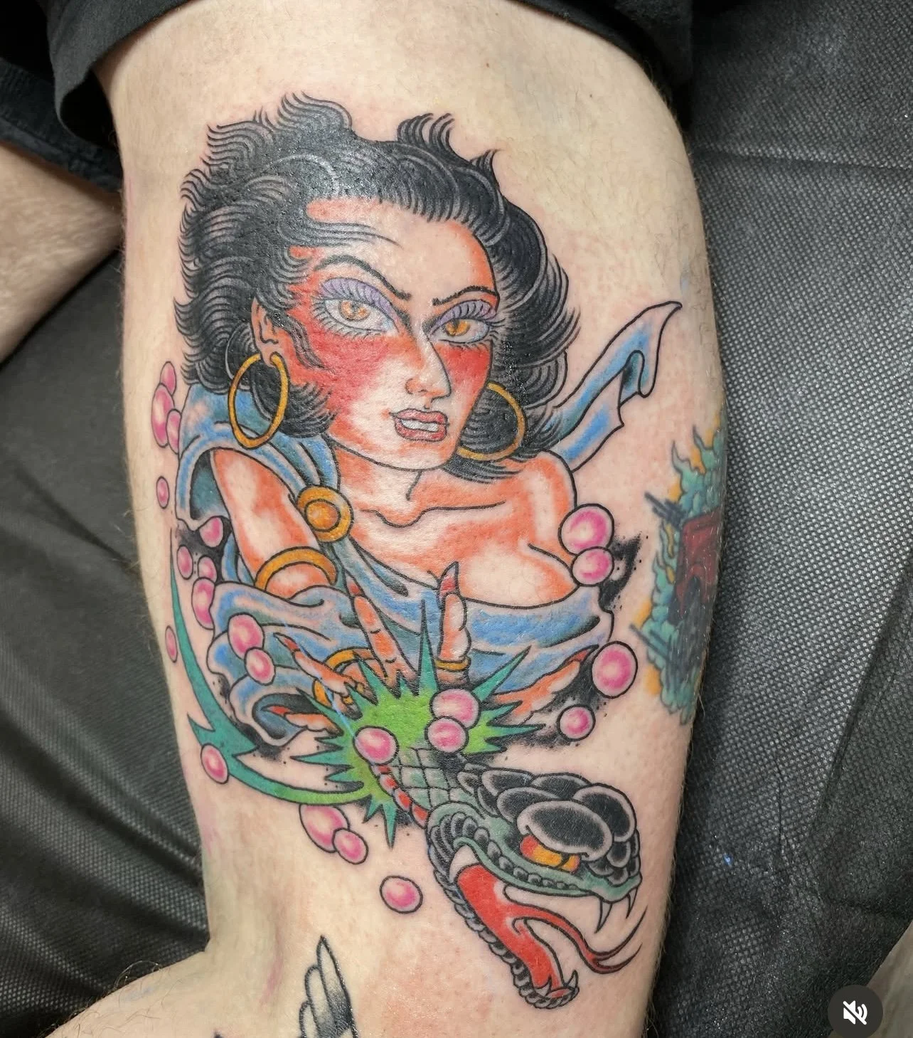 Colorful tattoo of a woman with dark hair, large earrings, and a blue dress, holding a dragon with pink beads and a green explosion.