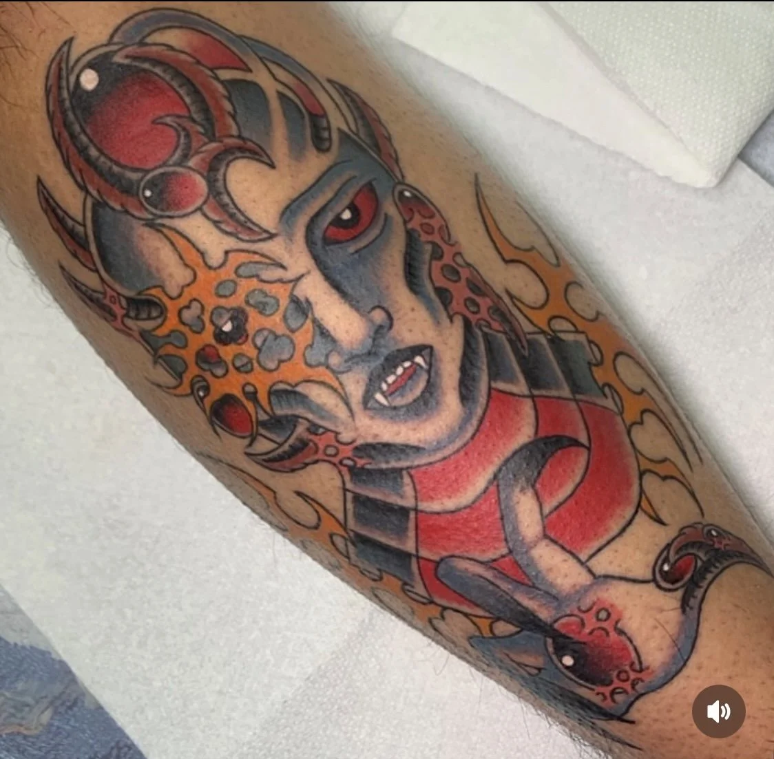 Tattoo of a fierce, stylized woman with red eyes and long curly hair, wearing a red top, surrounded by flames and leopard print patterns.