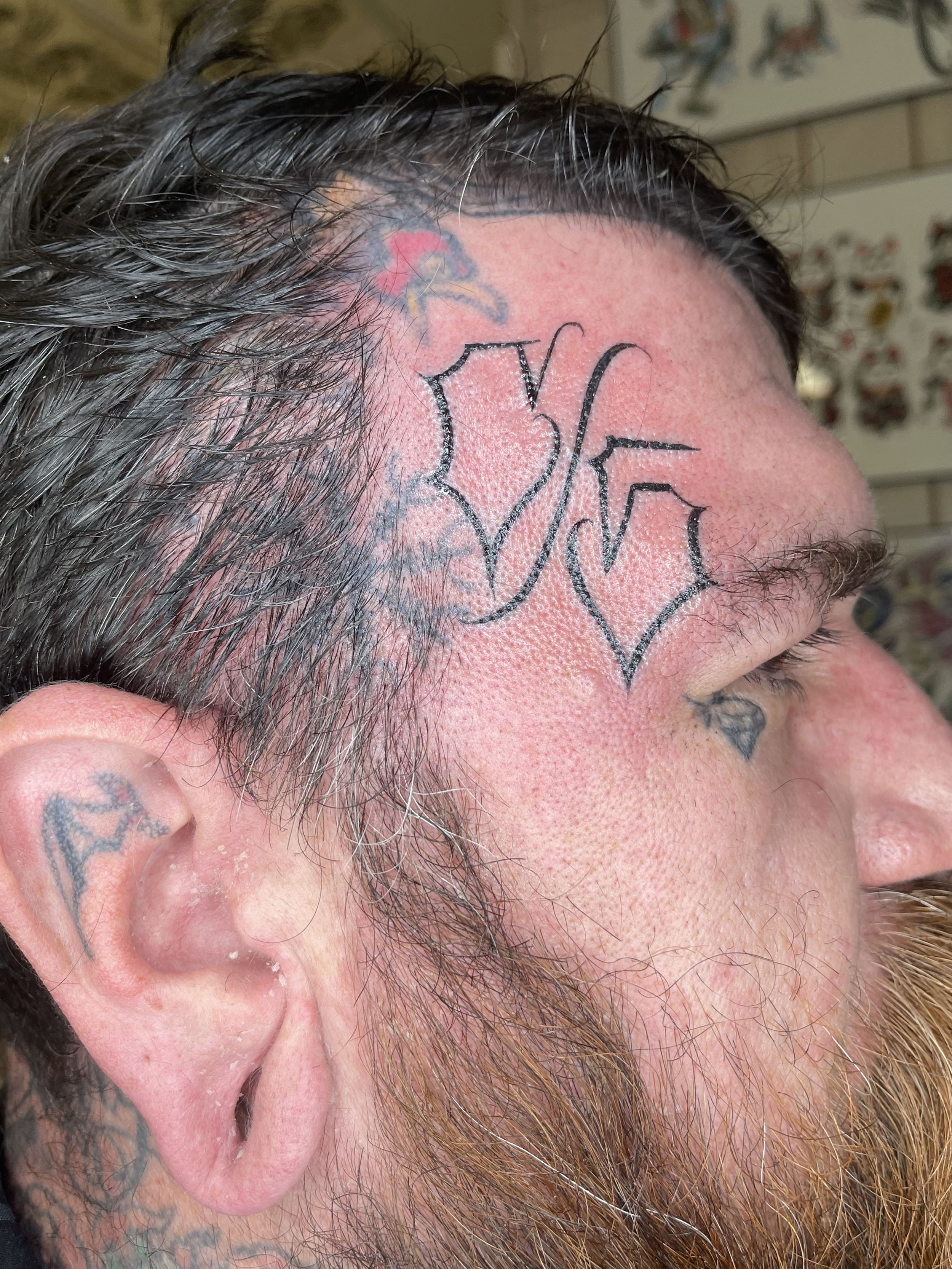 Close-up of a person's head showing short, dark, somewhat curly hair, tattoos on the scalp and behind the ear, and a beard on the side of the face.