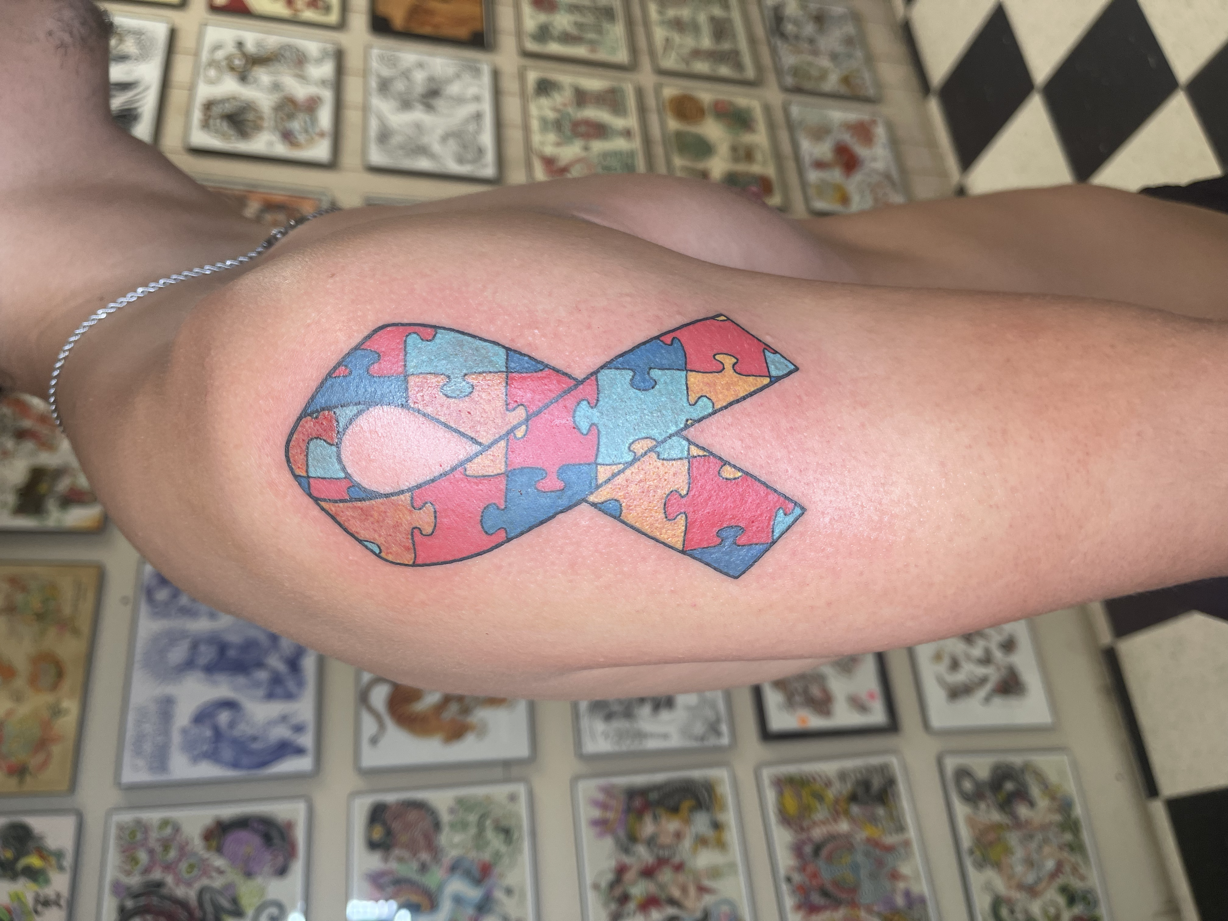 A tattoo of a heart filled with multicolored puzzle pieces on a person's upper arm. The background shows a wall with framed artwork and a black-and-white checkered floor.