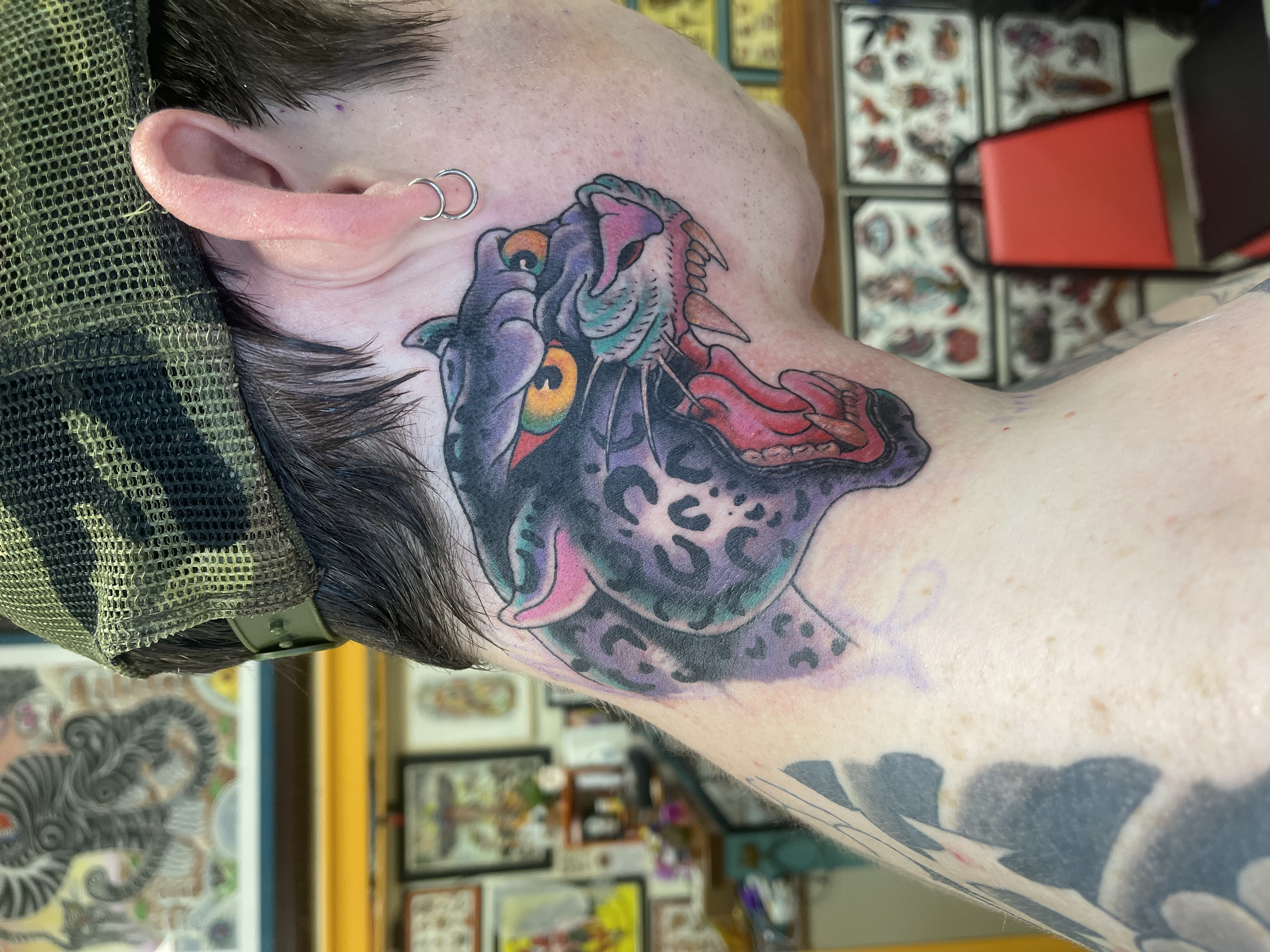 A person's shoulder and neck tattoo featuring a colorful dragon or snake with a rainbow eye, depicted in a vibrant, traditional style, in a tattoo shop background with tattoo flash and artwork on the walls.