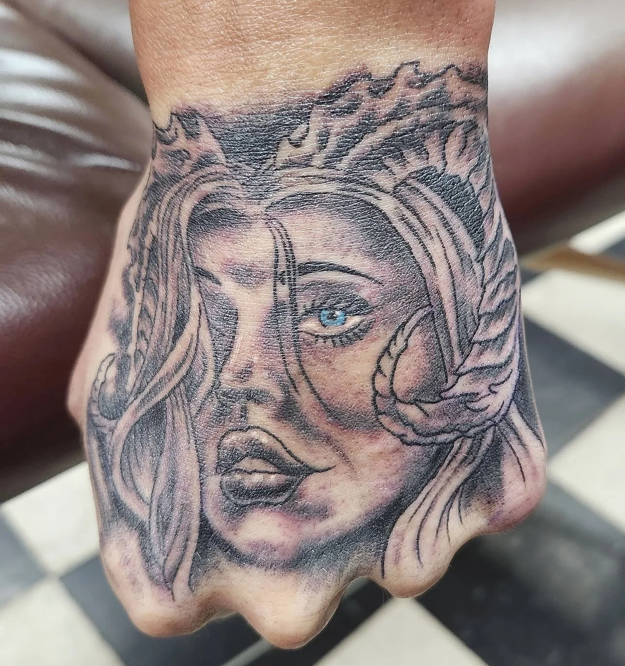 Close-up of a tattoo on a person's hand depicting a detailed portrait of a woman's face with long hair, piercing blue eye, and a slightly open mouth.