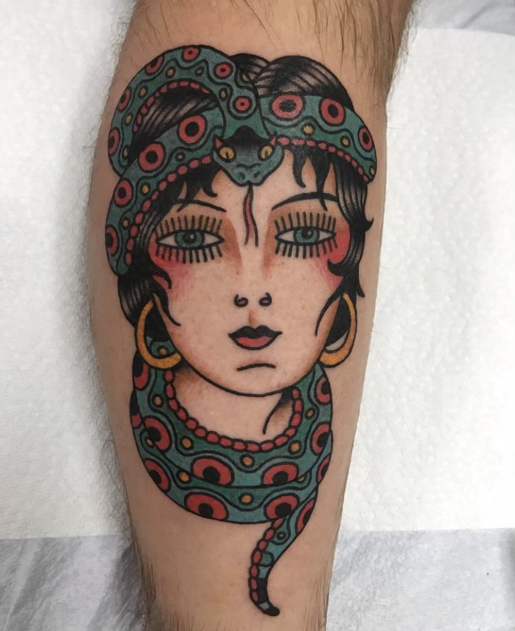 Tattoo of a woman’s face with a snake wrapped around her head and neck, featuring large eyes, gold hoop earrings, and intricate colorful patterns.