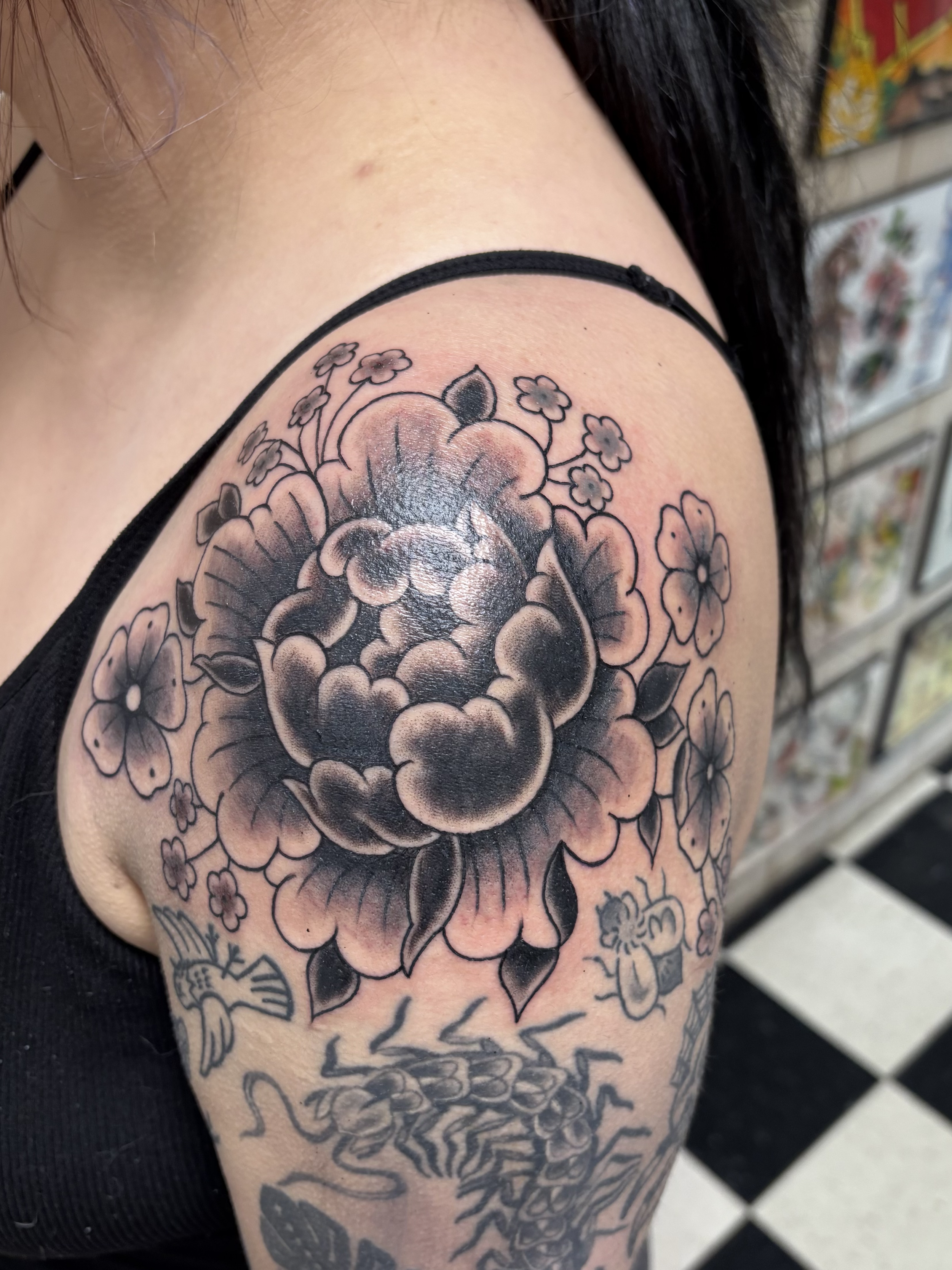 Close-up of a person's upper arm with a large black and gray floral tattoo, featuring a central flower, smaller surrounding flowers, leaves, and some abstract elements.