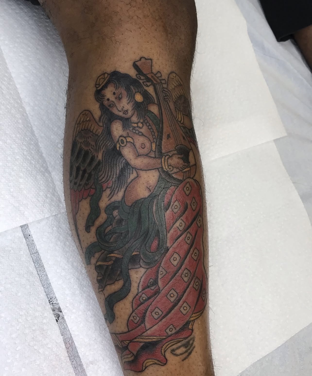 Tattoo of a winged woman with long black hair, playing a stringed instrument, surrounded by flowing fabric, on a person's leg.