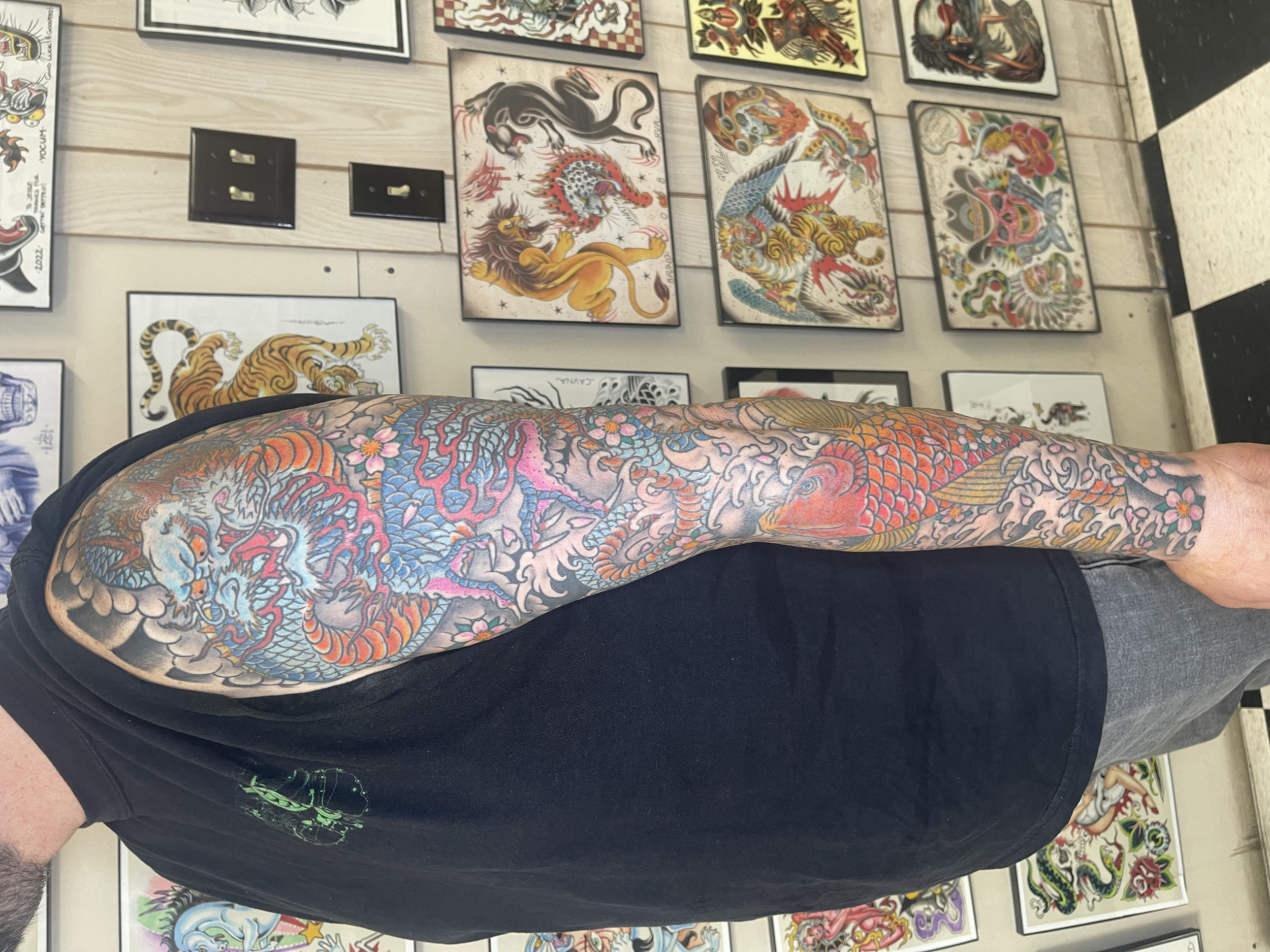 Close-up of an arm covered in vibrant traditional-style tattoo featuring a dragon surrounded by flowers and clouds, with colorful detailed artwork on the tattoos.