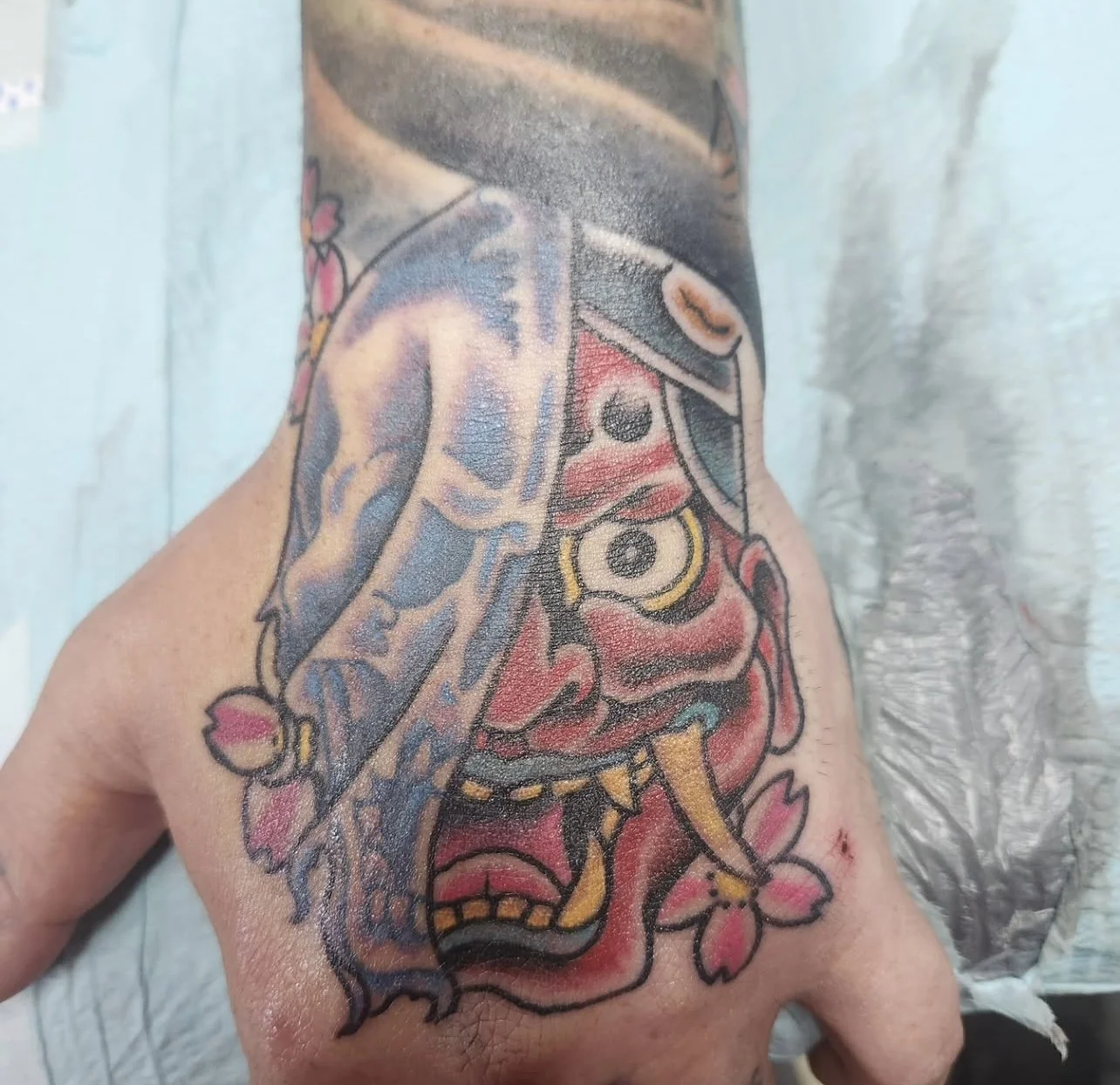 Close-up view of a colorful tattoo on a person's arm, featuring a traditional Japanese demon mask with a red face, large eyes, protruding yellow tongue, and decorated with cherry blossoms.