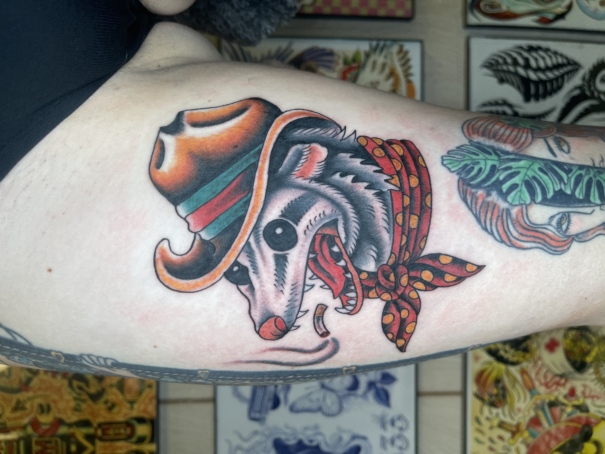 Colorful tattoo of a dog's skull wearing a spacesuit and helmet, with green leaves and orange accents, on a person's arm, with tattoo flash art in the background.