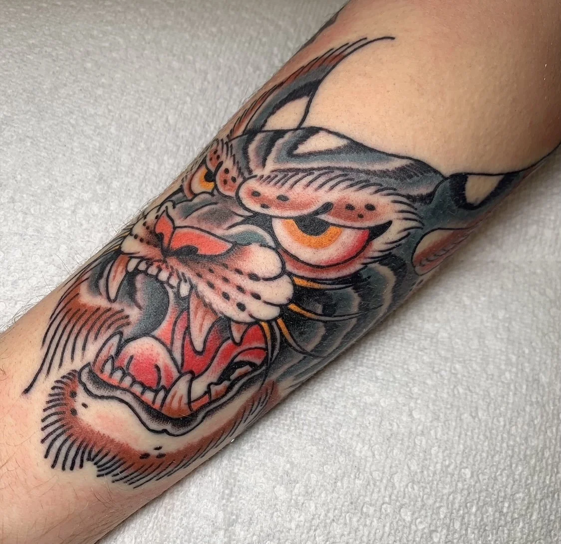 Tattoo of a fierce tiger face with orange eyes, open mouth revealing sharp teeth, and prominent whiskers on a person's arm.