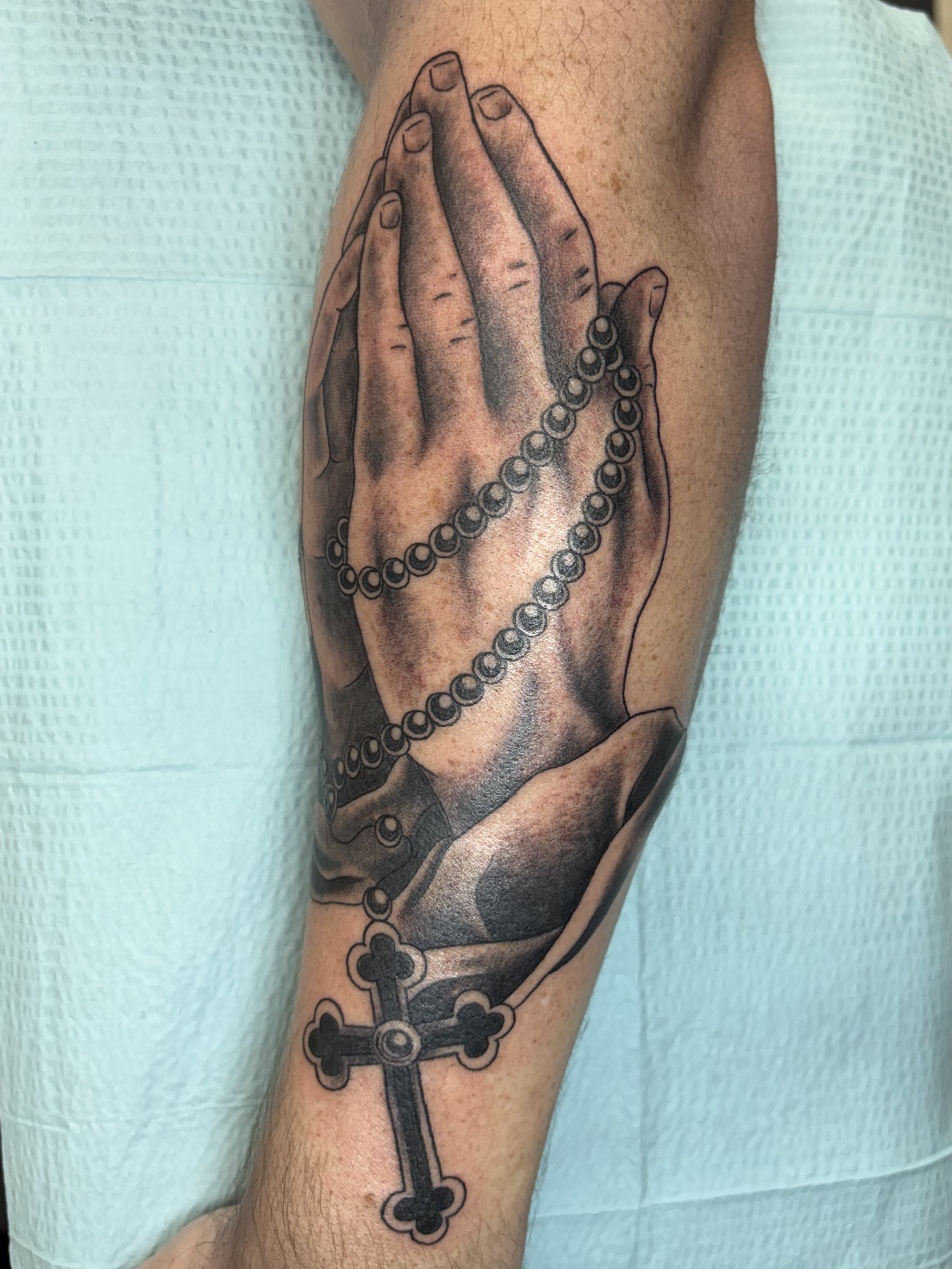 Tattoo of praying hands holding rosary beads on person's forearm.