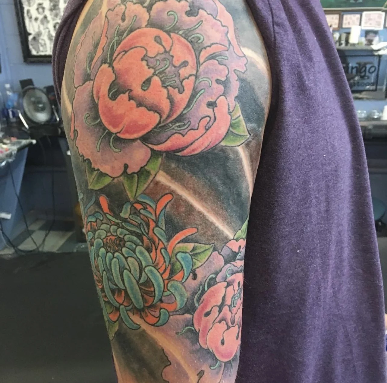Tattoo of a large pink peony flower with green leaves and orange, green, and blue chrysanthemums on a person's upper arm.