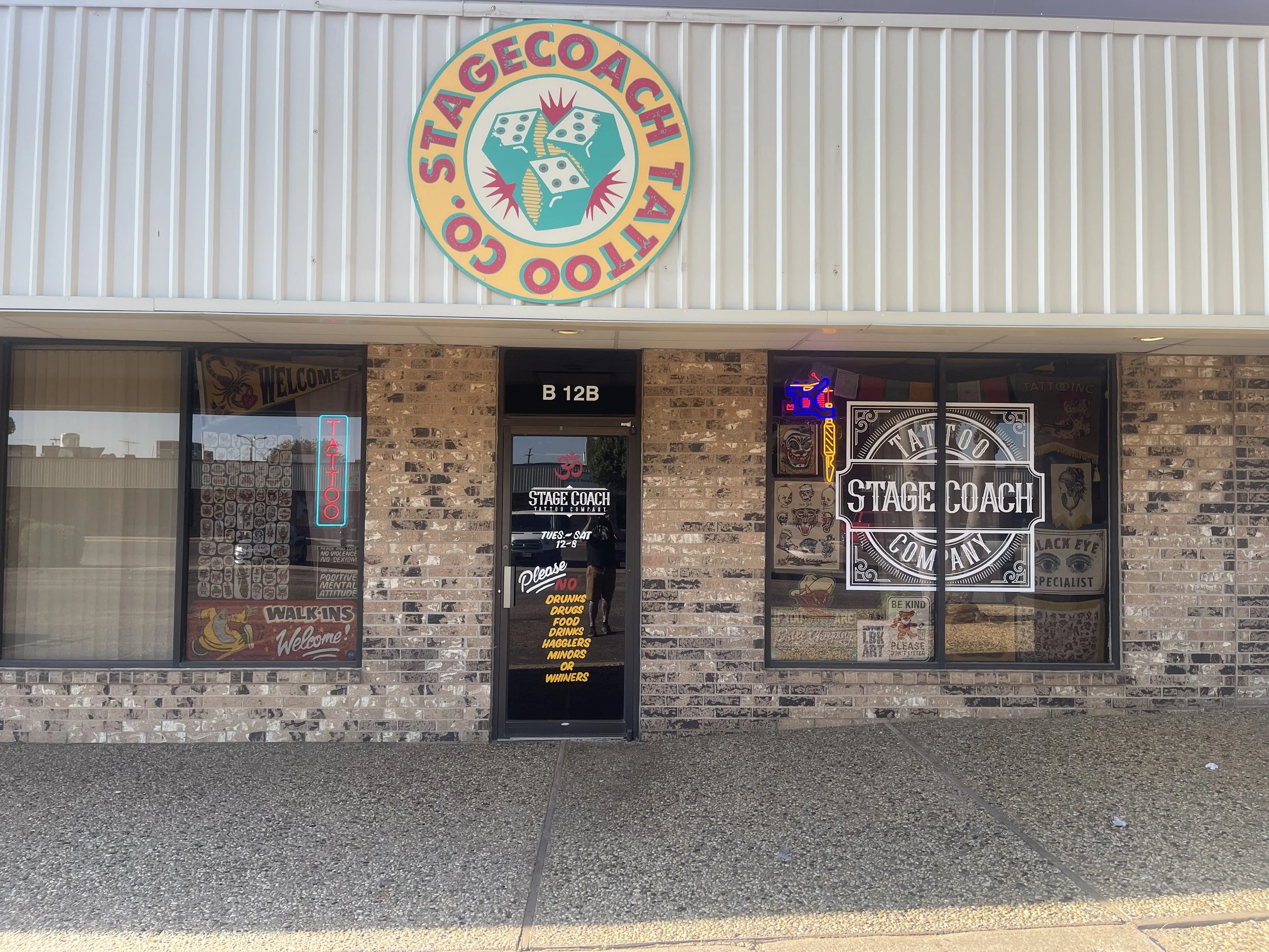 Storefront with a circular sign that reads "STAGE COACH TATTOO CO" with a logo of a stagecoach, and black brick walls with windows showcasing tattoo art, neon signs, and notices.