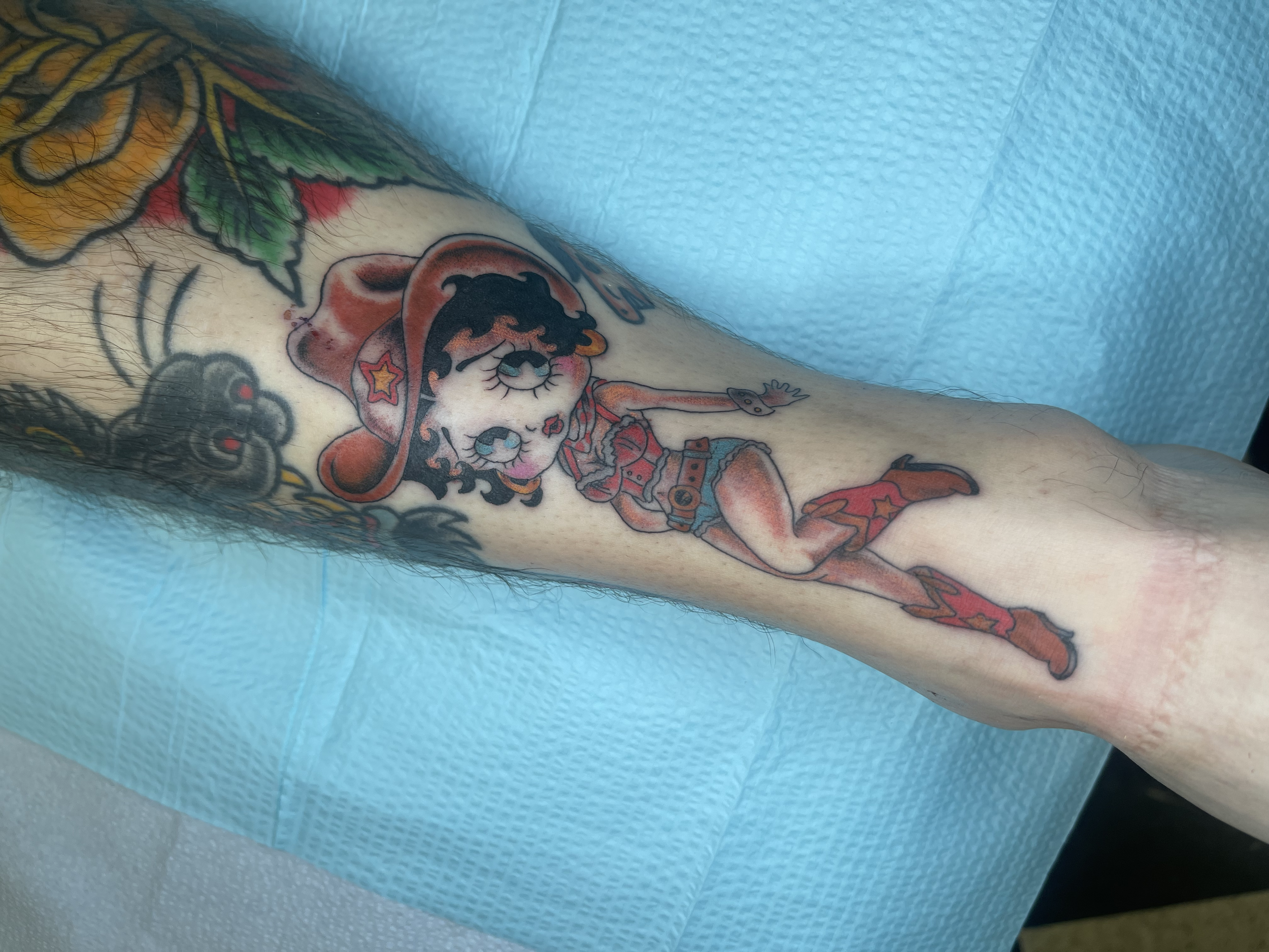 Tattooed arm with colorful cartoon-style tattoos, including a girl with big eyes and a Santa hat, a green leaf, and other designs, on a blue textured surface.