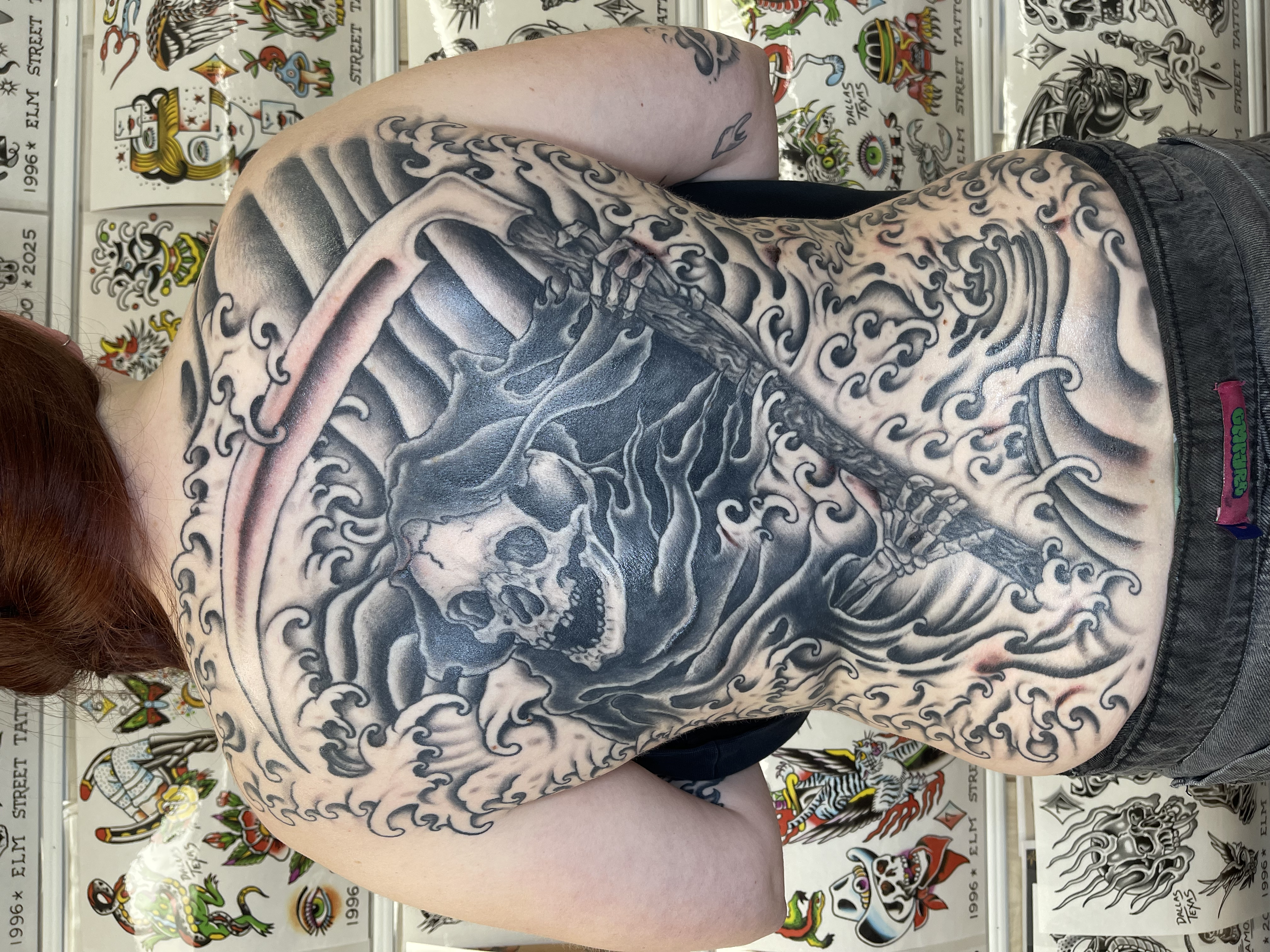 A person's back and arm with a detailed black and gray tattoo of a skull and swirling wave patterns, with a background of tattoo flash artwork on a wall.