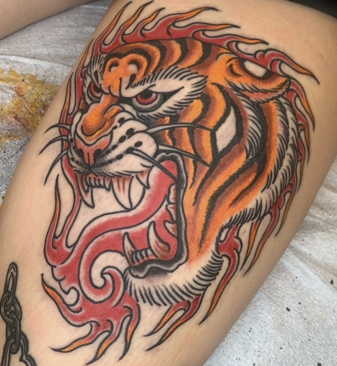 A colorful tattoo of a roaring tiger head with orange, black, white, and red details, including flames around its head.