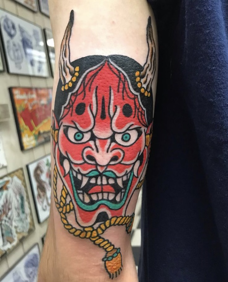 Colorful traditional Japanese demon mask tattoo on arm with red, black, teal, and yellow details.