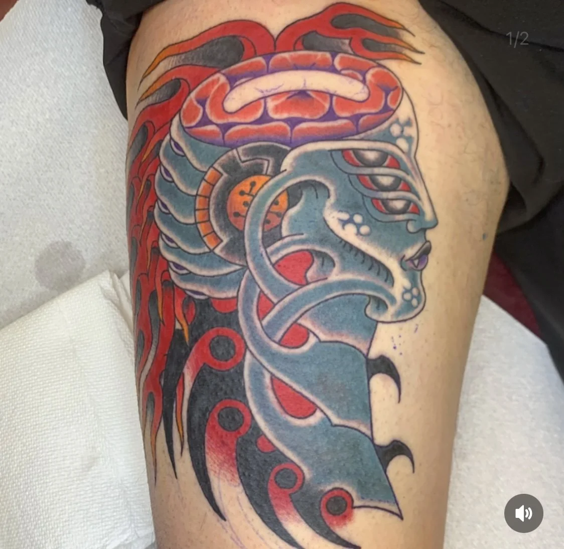 A colorful tattoo on a person's thigh featuring a stylized female face with closed eyes, surrounded by abstract and organic shapes in shades of blue, red, orange, and black.