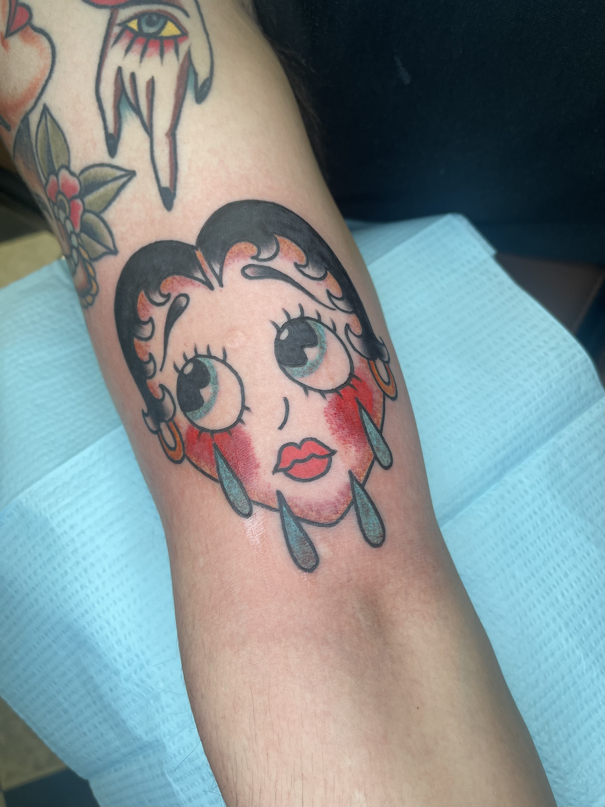 Tattoo of a stylized woman's face with large eyes, bold lips, and earrings, outlined in black with orange and teal accents, on a person's arm.