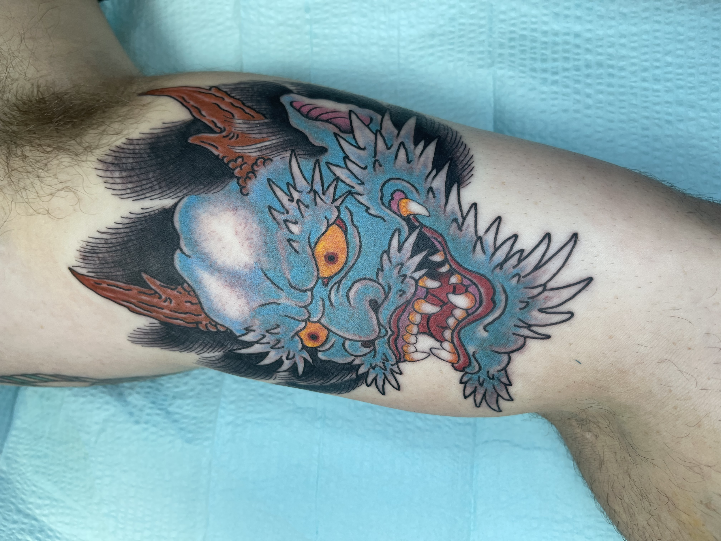 A colorful dragon tattoo on a person's upper arm, featuring blue, orange, pink, and white colors with detailed line work and shading.