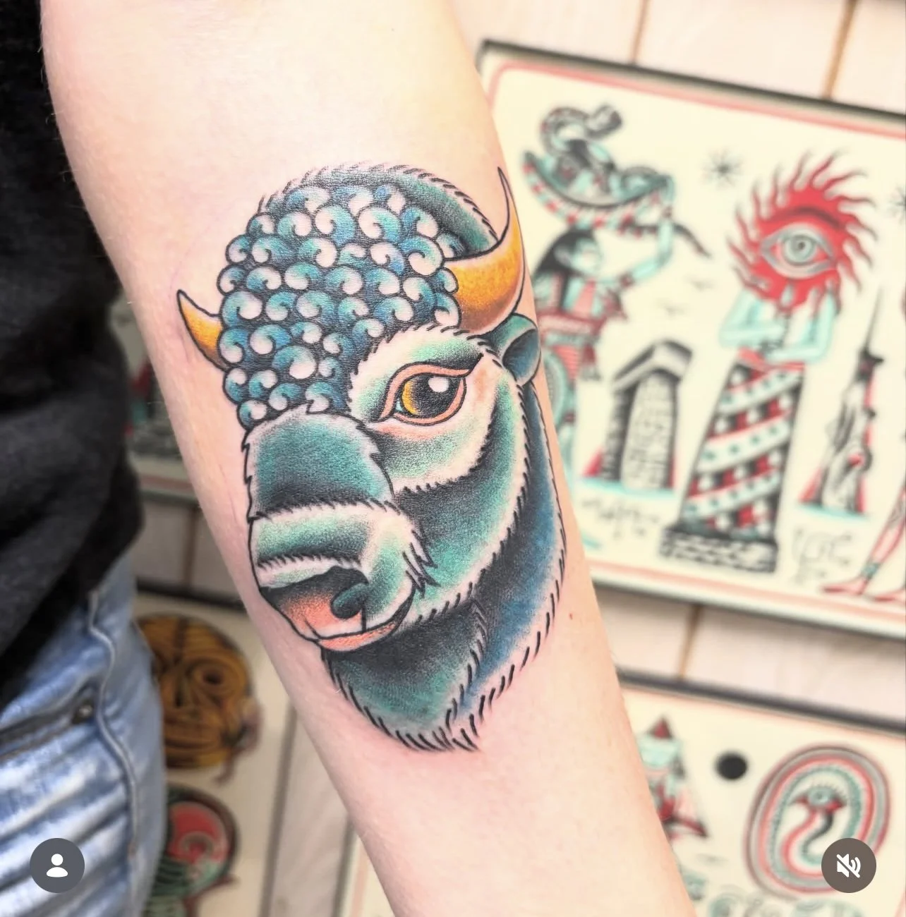 Colorful tattoo of a bison's head with curly hair and horns on an arm. Background features various tattoo art designs.