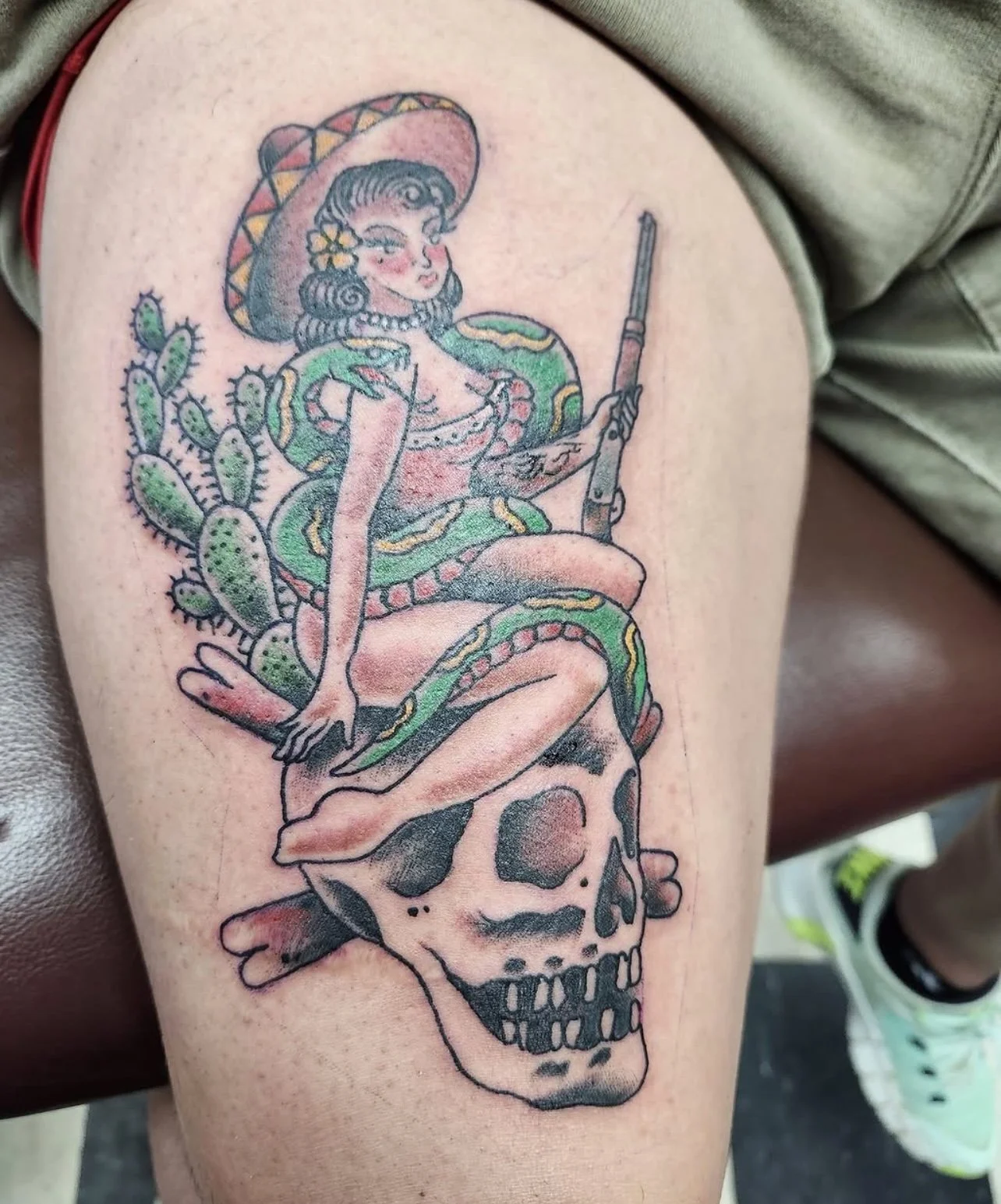 Tattoo of a woman sitting on a skull with a snake wrapped around her, holding a rifle, with cacti in the background, in a traditional tattoo style.