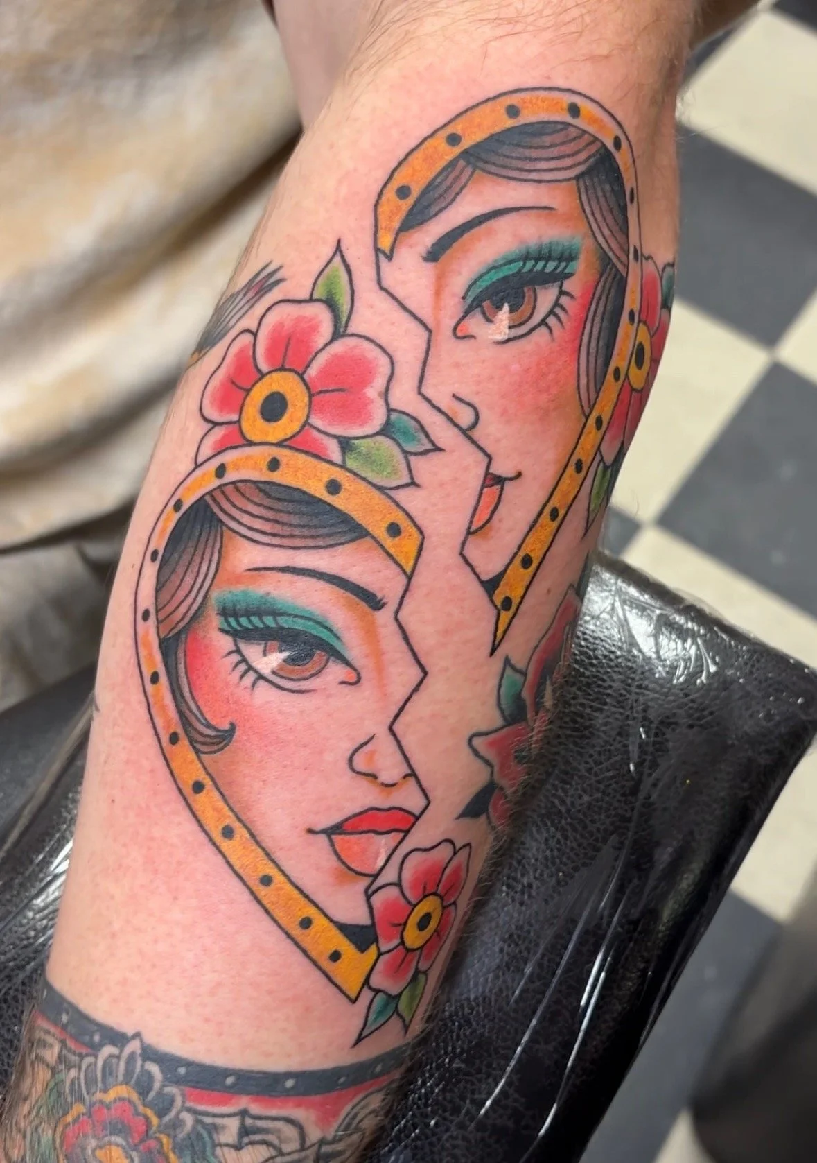 Tattoo of two faces with matching features, separated by a crack, surrounded by colorful flowers on a person's arm.