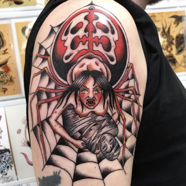 Tattoo of a woman with long black hair sitting in a spider web, holding a knife and a mask, with a large spider above her and a skull symbol in the background.