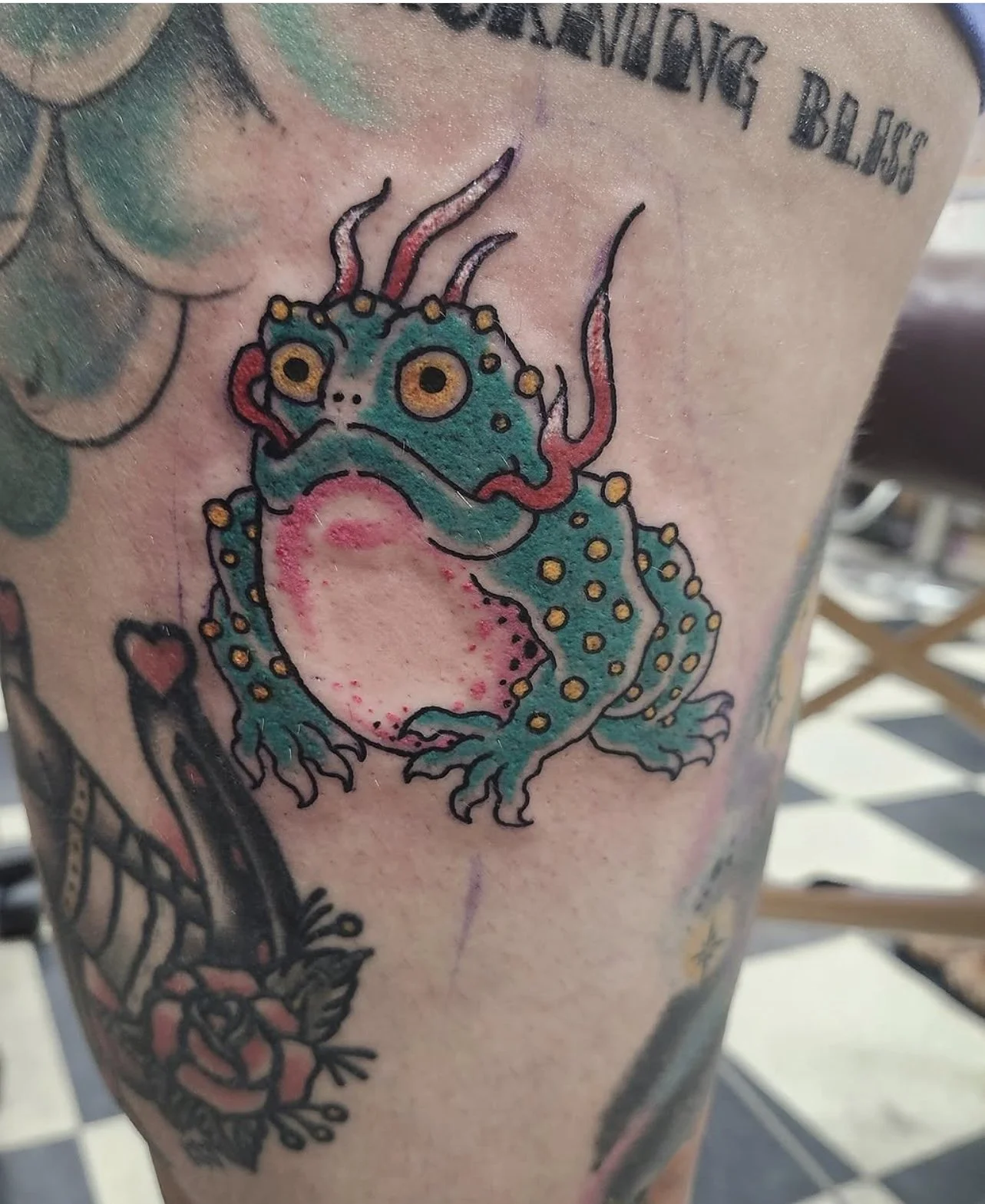 Tattoo of a colorful, cartoon-style frog with large yellow eyes, red tongue, and yellow spots on its teal body, on person's skin.