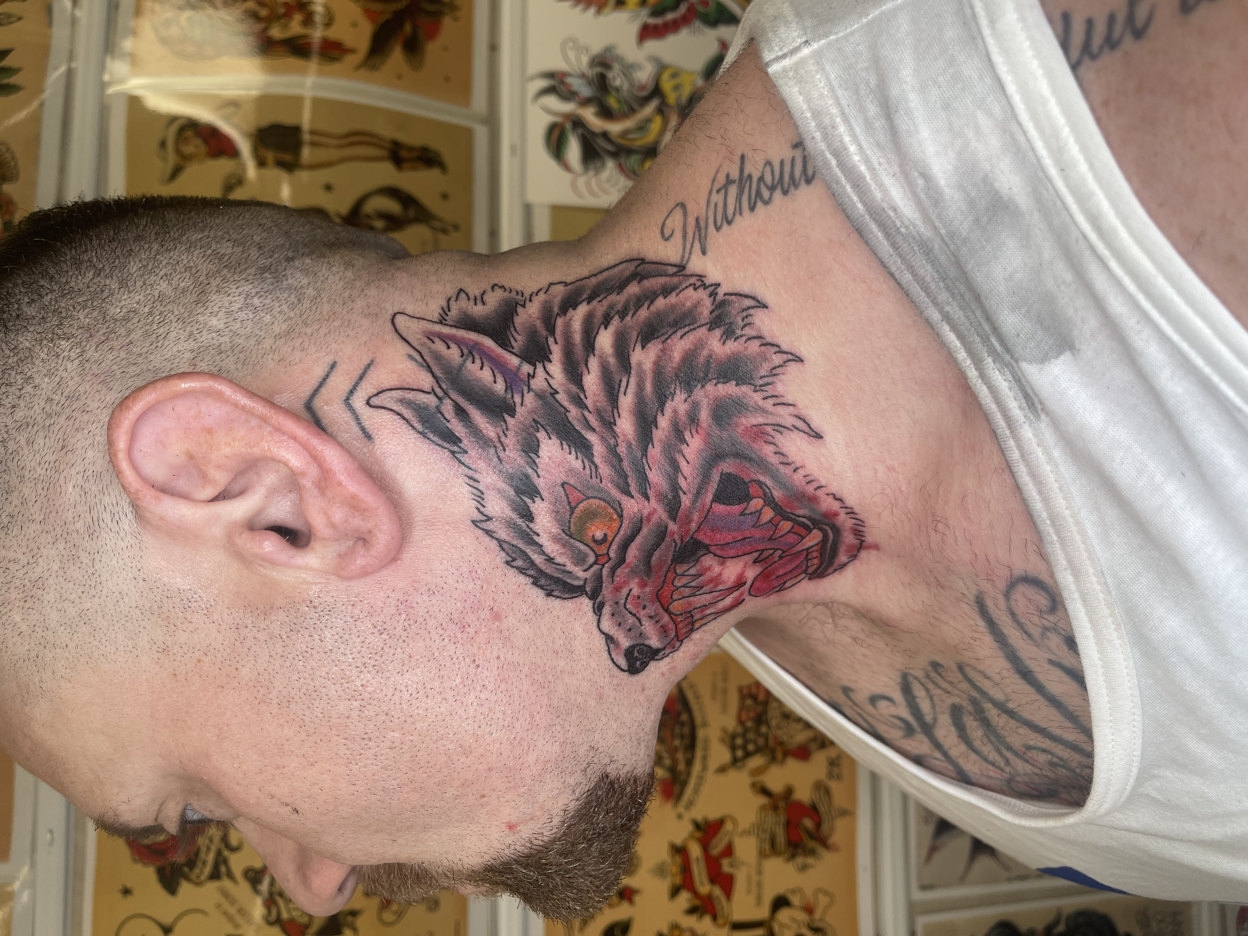 Close-up of a man's side face with a tattoo of a wolf or fox on his neck. The tattoo is detailed with dark shading and colorful eyes. The man has a short hairstyle, a beard, and is wearing a white shirt. The background contains colorful illustrations