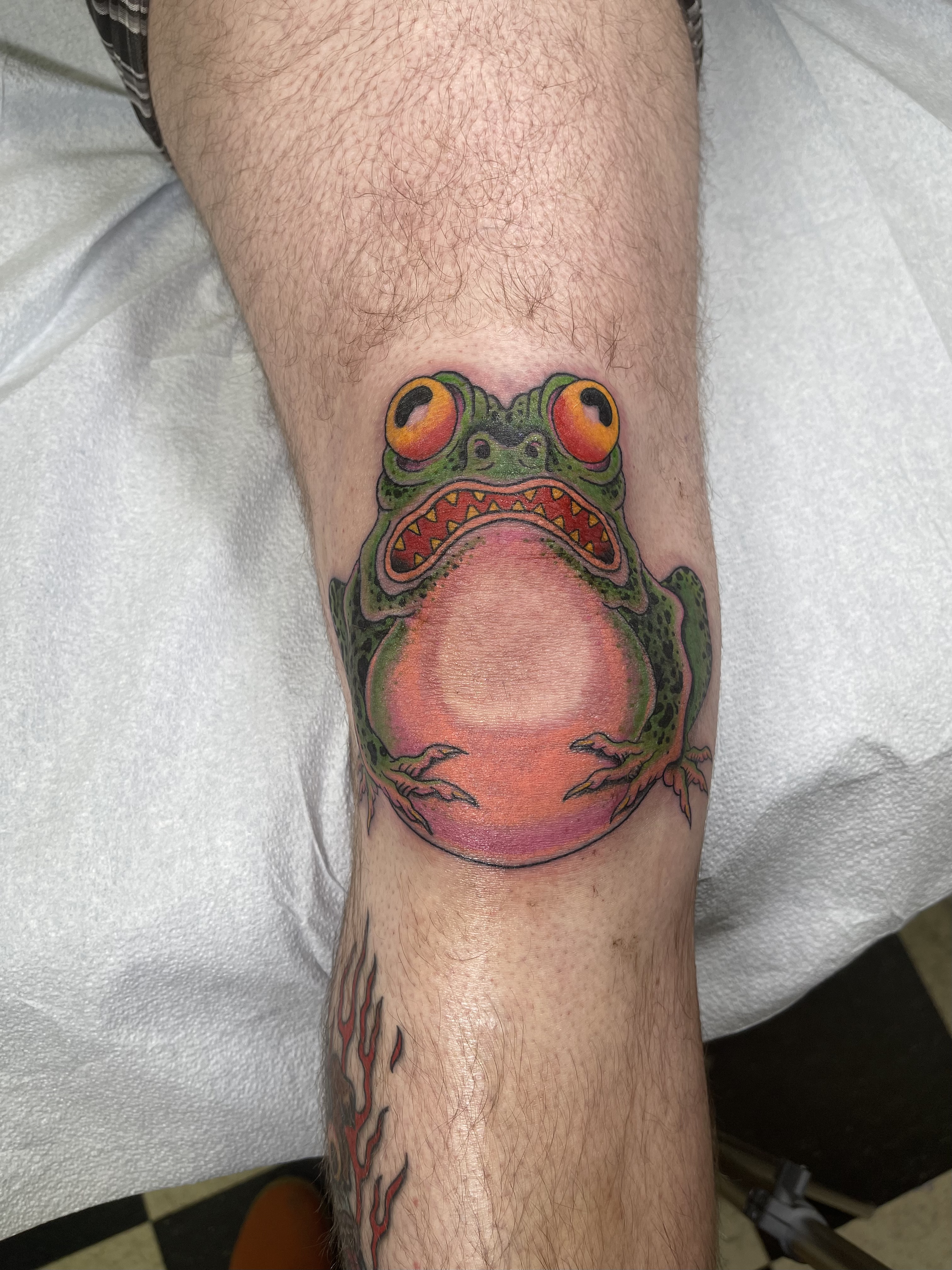 Colorful tattoo of a frog on a person's leg, with large orange and black eyes and an open mouth, sitting on a pinkish background with red and black accents.
