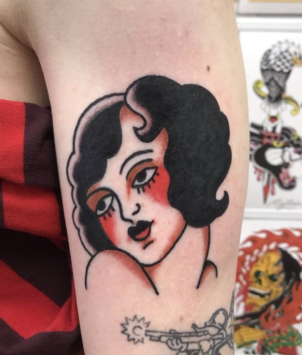 Tattoo of a stylized woman with dark curly hair and red blush on her cheeks, holding a tattoo machine.