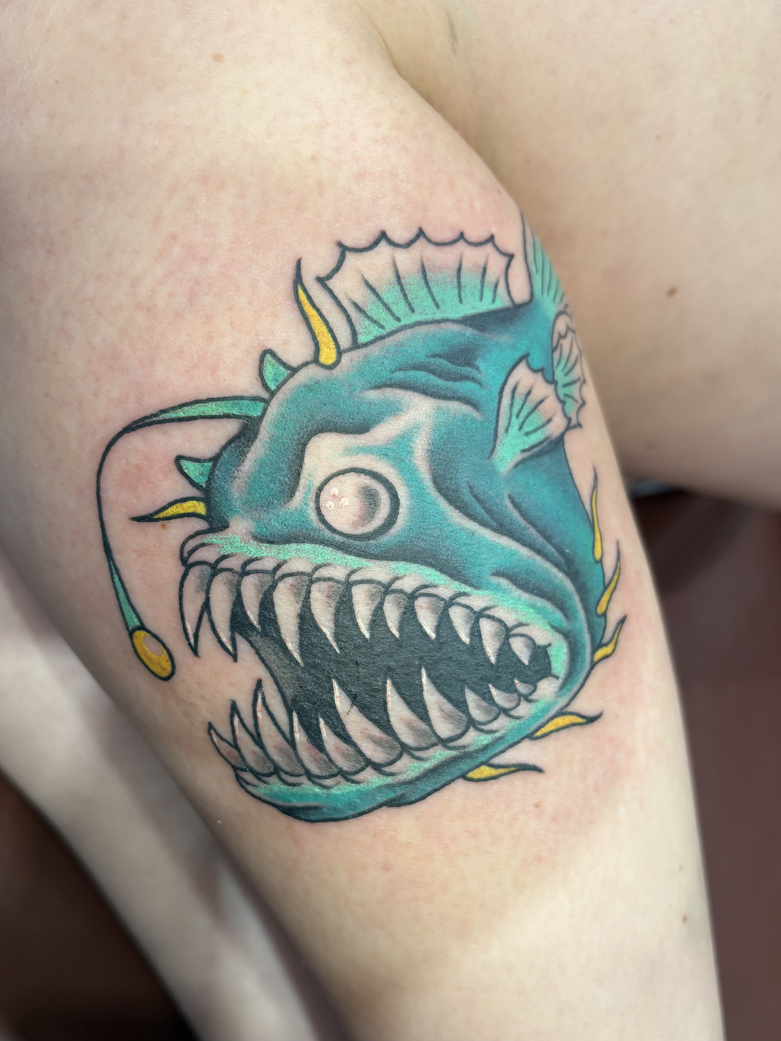 A tattoo on the person's shoulder of a fish skull with sharp teeth, colored in shades of teal, blue, yellow, and black.
