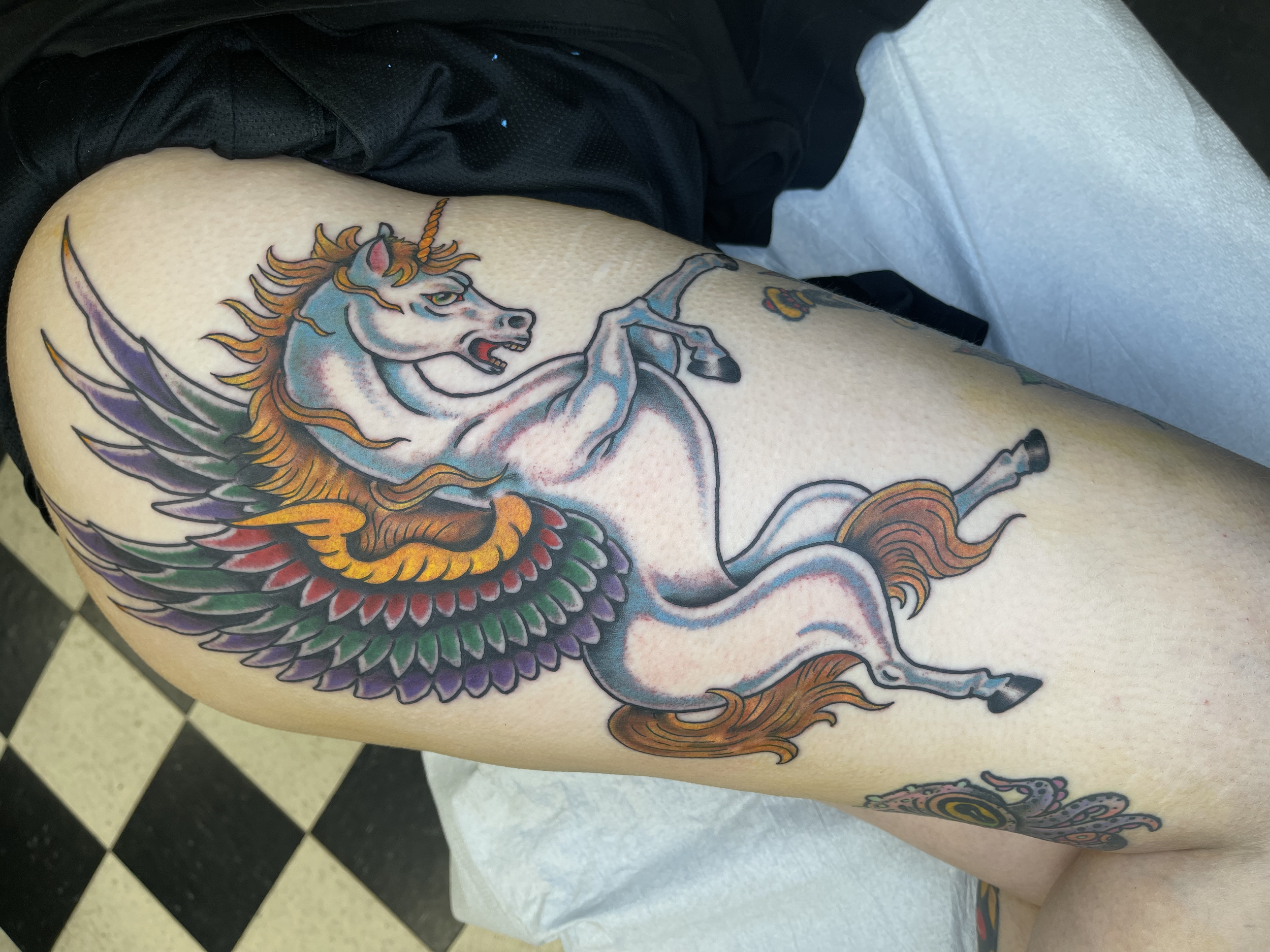 Colorful tattoo of a unicorn with wings on a person's leg, with a black and white checkered floor in the background.Lubbock tattoo artists Stagecoach Tattoo Co.