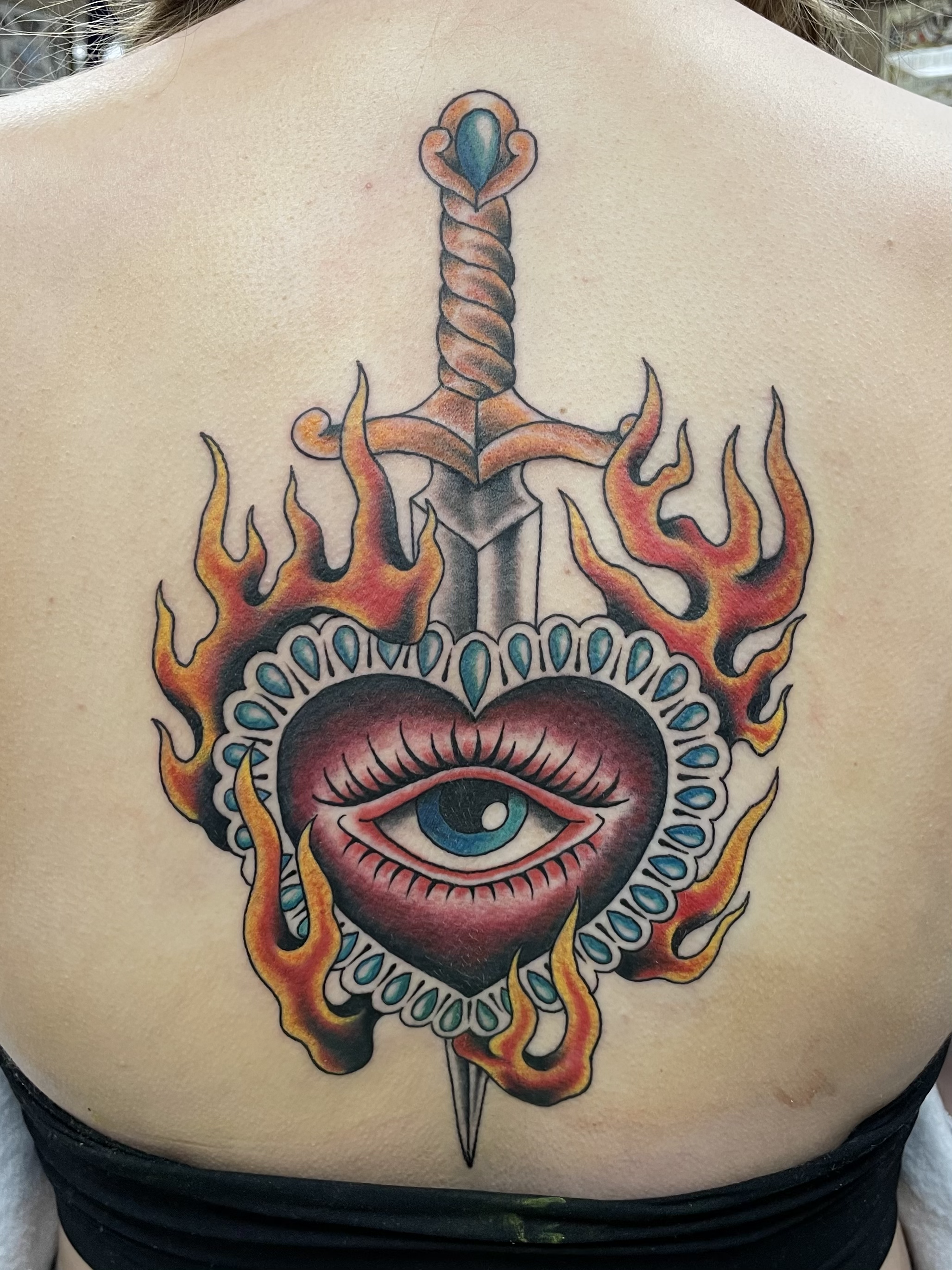 A tattoo on a person's chest featuring an eye within a heart, surrounded by flames, with a large sword piercing through the design.