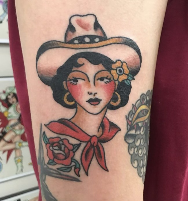 A tattoo of a woman with vintage style, wearing a wide-brimmed hat decorated with a heart and flowers, red scarf, and large gold hoop earrings, with traditional tattoo style roses and a skull in the background.