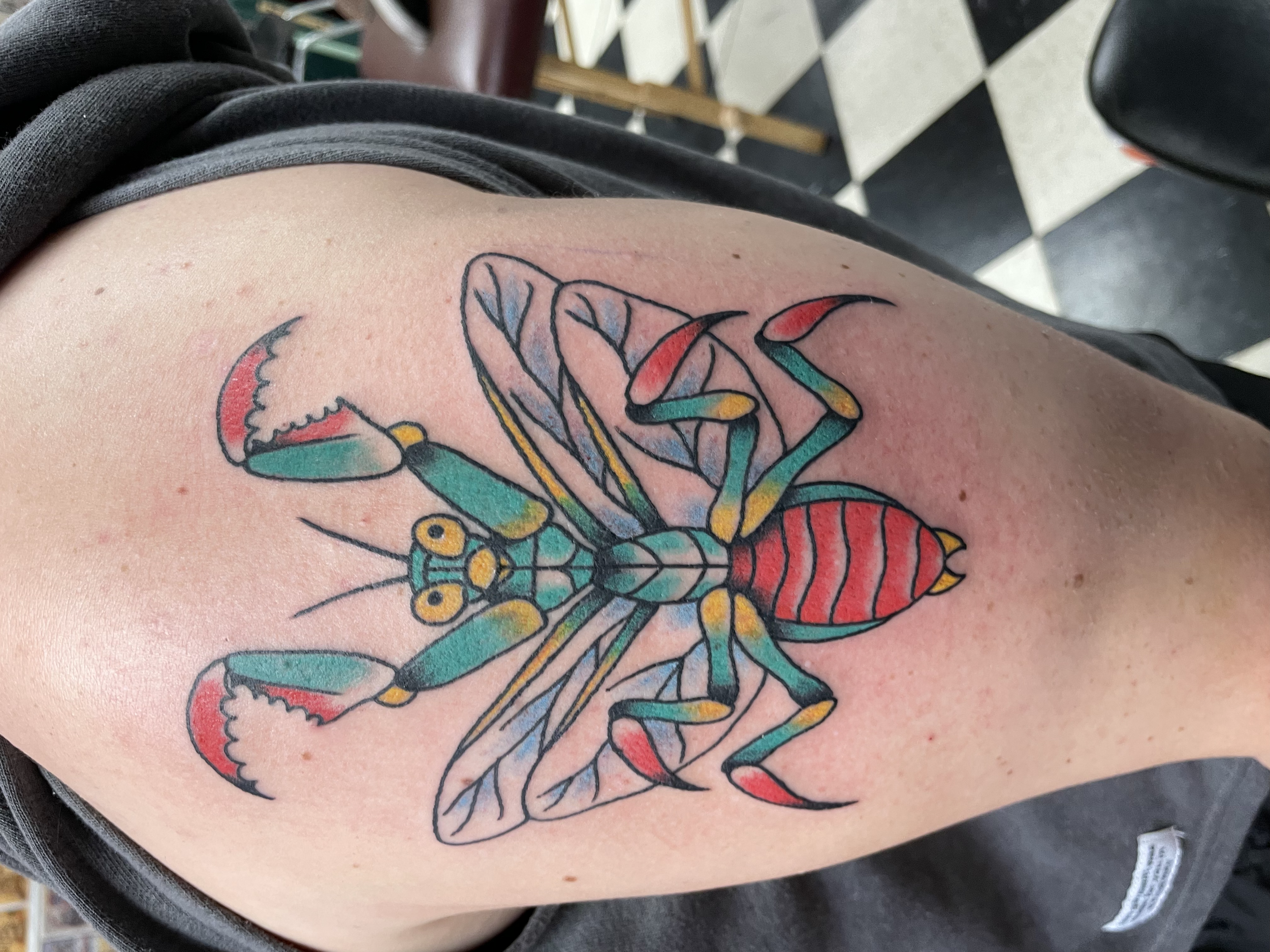 Colorful tattoo of a curled lobster on a person's upper arm with a background of black and white checkered flooring.
