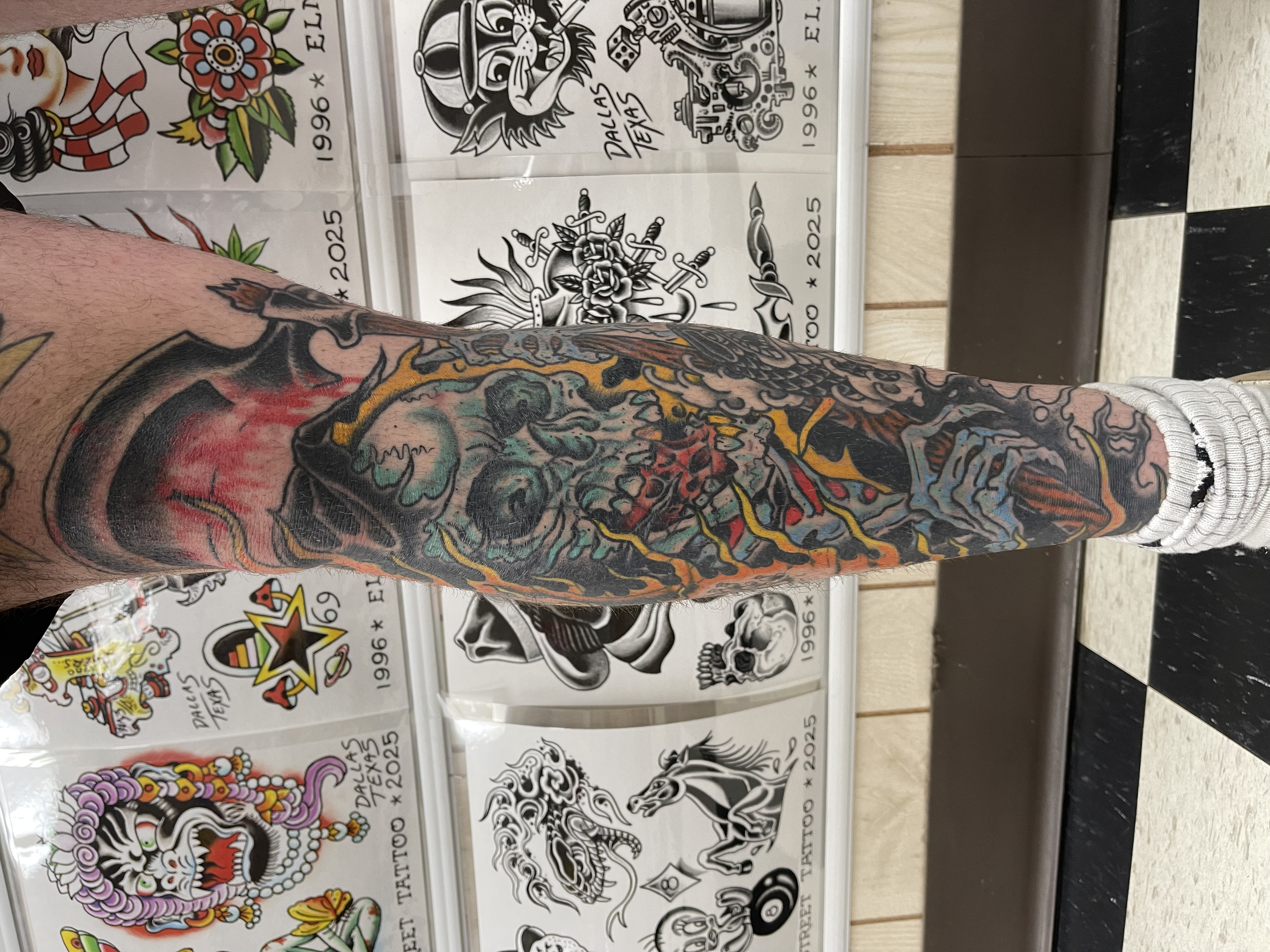 Tattoo designs on a person's arm and tattoo flash sheets in the background with various tattoo flash art and lettering.