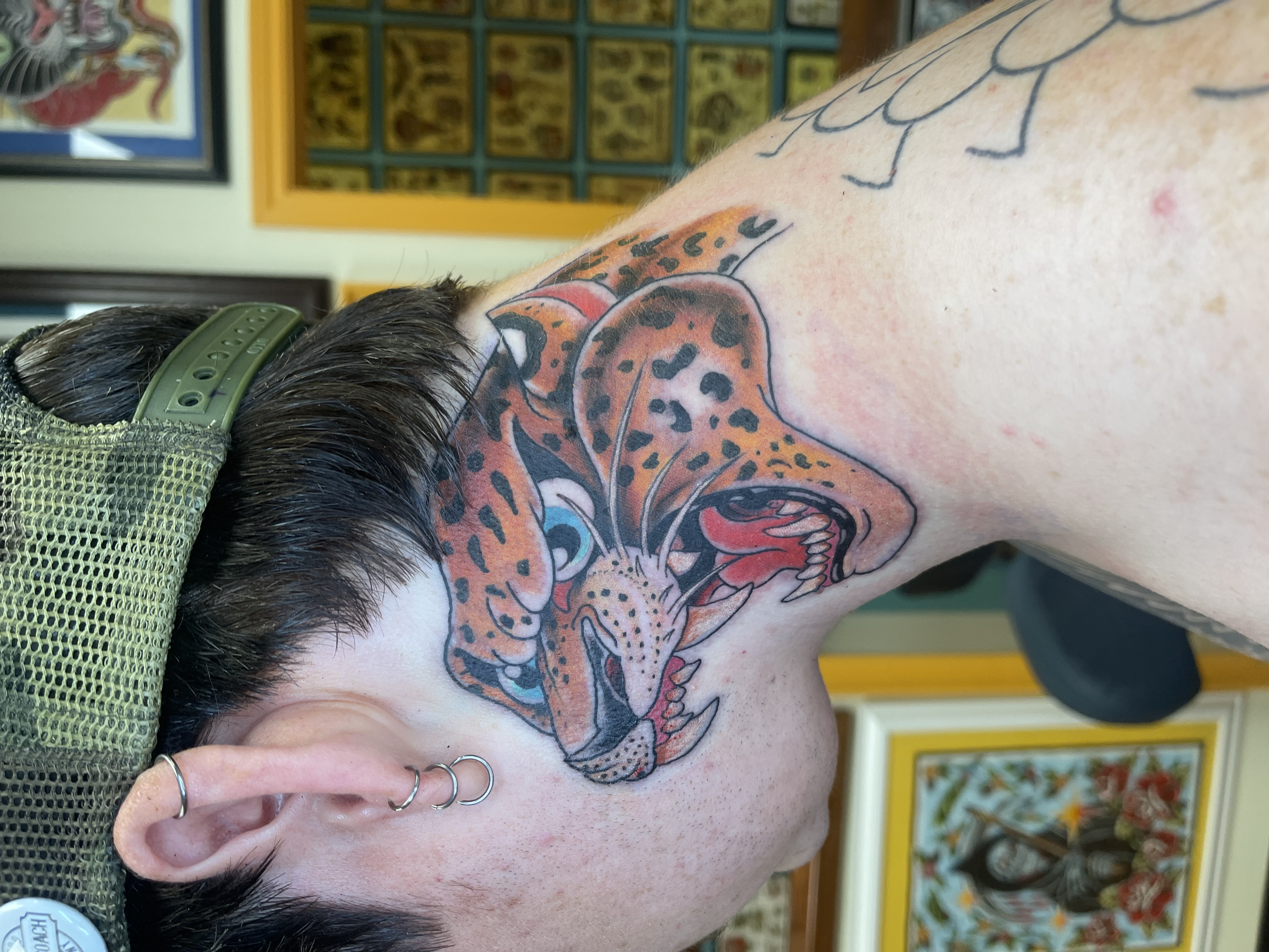 Close-up of a person's neck with a colorful tattoo of a cheetah or leopard head, dark hair, and multiple piercings on the ear, in an indoor setting with framed artwork on the wall.