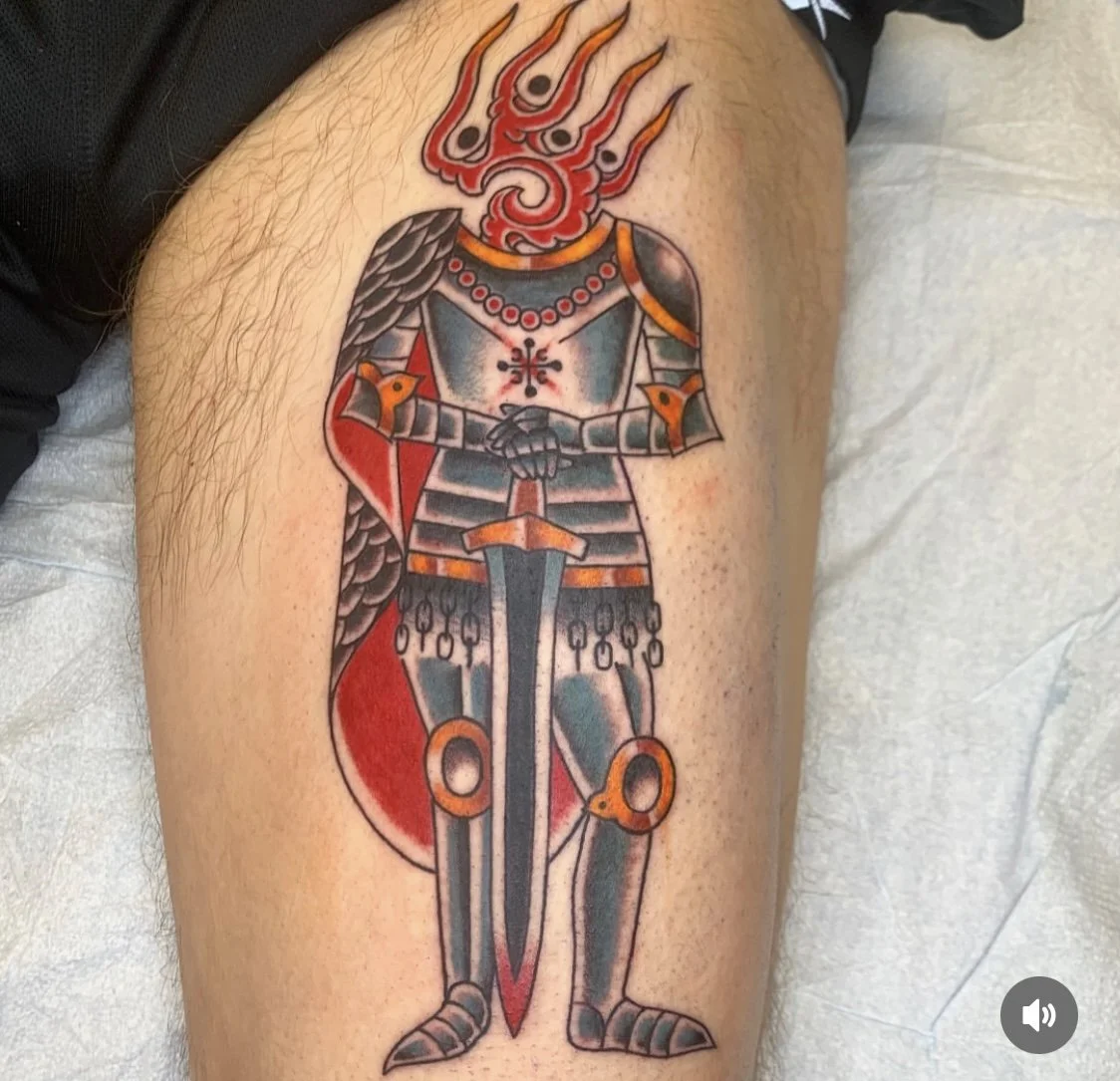 Tattoo of a knight in armor with a fiery dragon head for a helmet, holding a sword, with a red shield and a red cape.