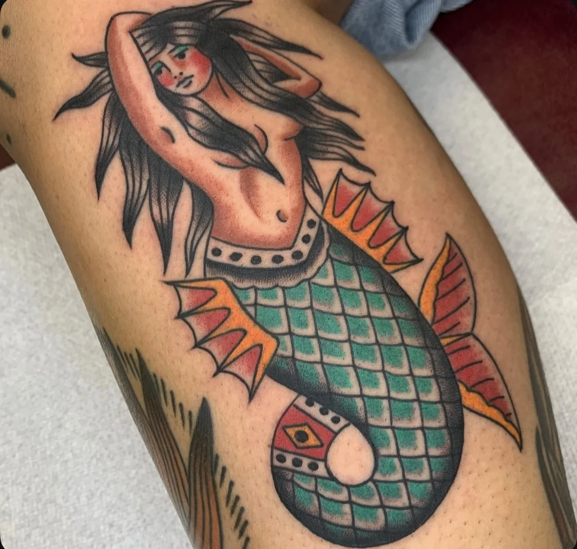 Tattoo of a mermaid with long black hair, a human upper body, and a fish tail with scales in shades of blue and green, with fins in orange and yellow, on a person's arm.