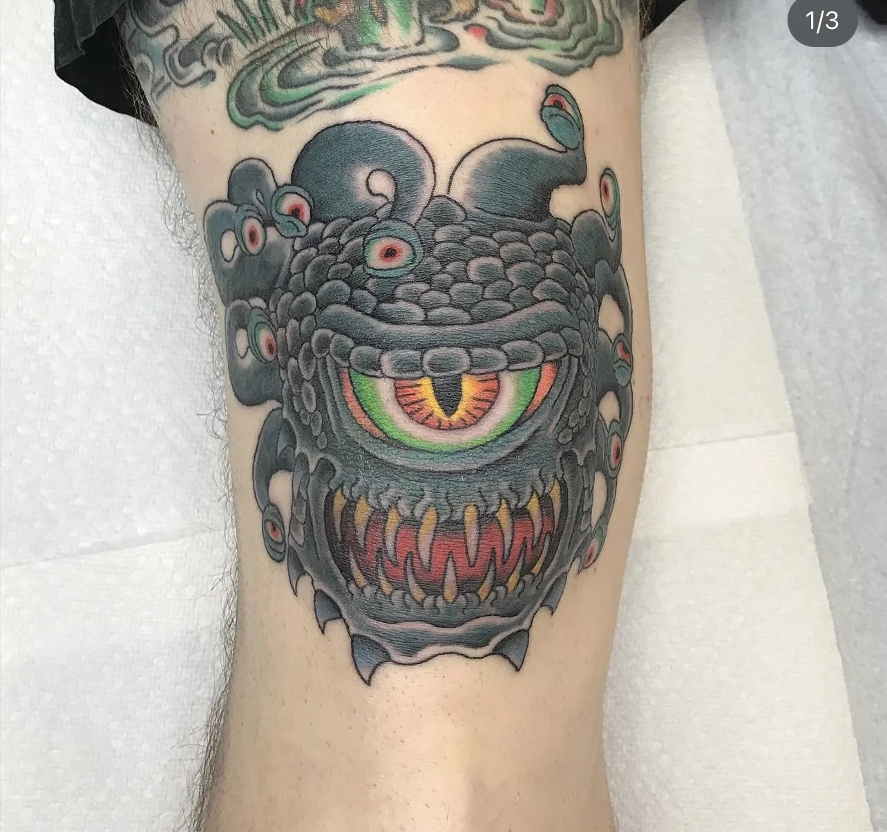 Colorful tattoo on leg featuring a monstrous creature with a large eye in the center, surrounded by tentacle-like appendages and sharp teeth, with vivid colors.