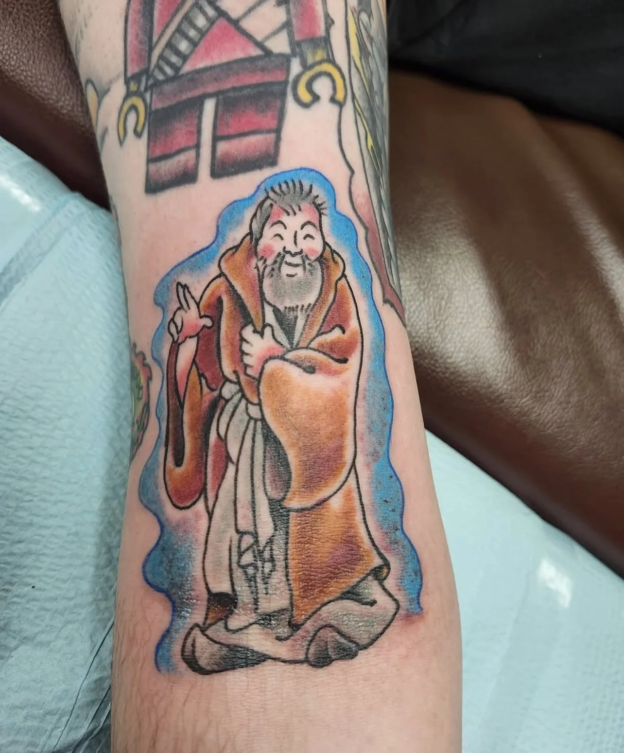 Colorful tattoo of a smiling, bearded man dressed as a monk or sage, making a peace sign with his right hand, on a person's forearm.