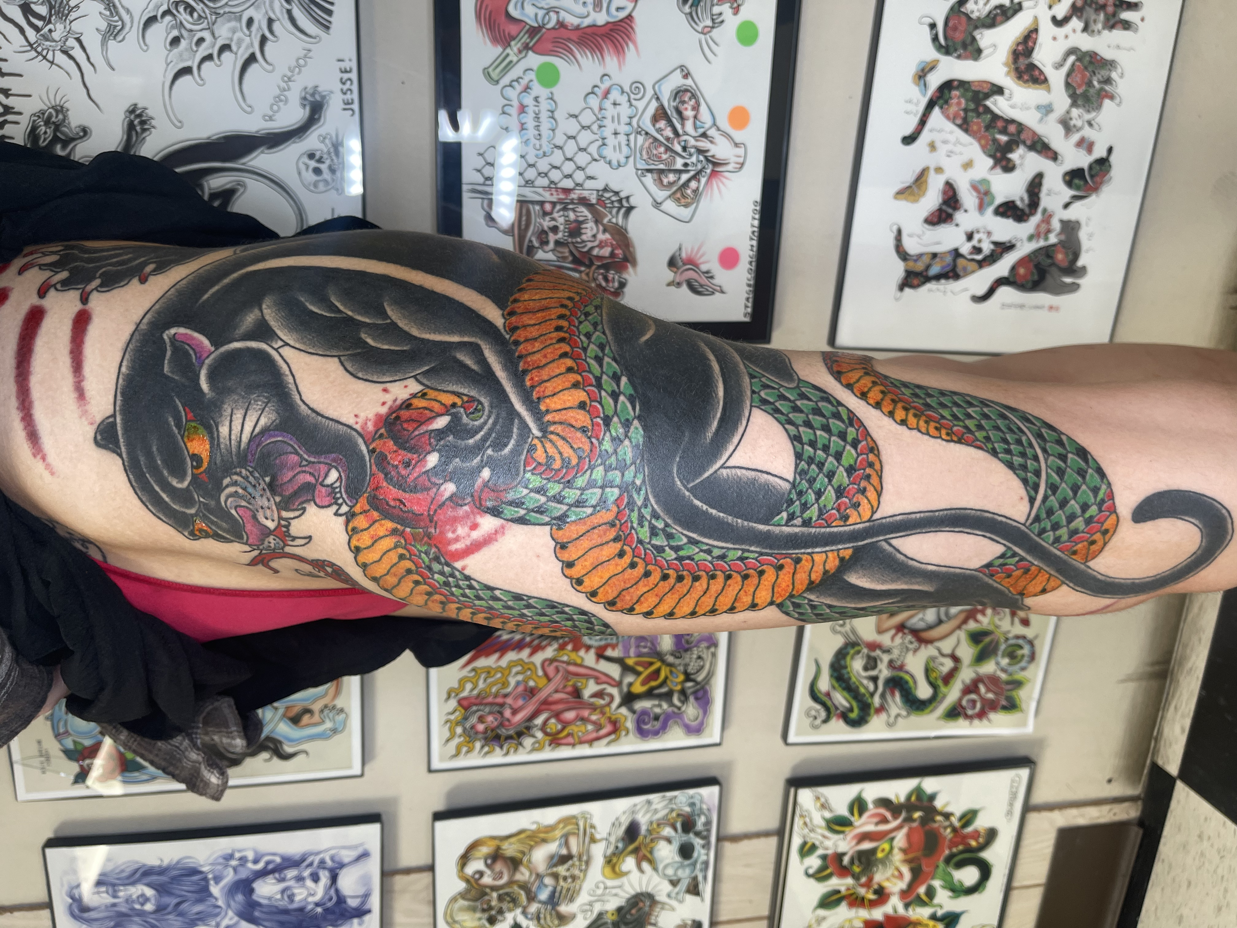 Tattoo artist displaying traditional Japanese style tattoo designs on sheets of paper, with a full sleeve tattoo featuring a dragon on a person's arm in the foreground.